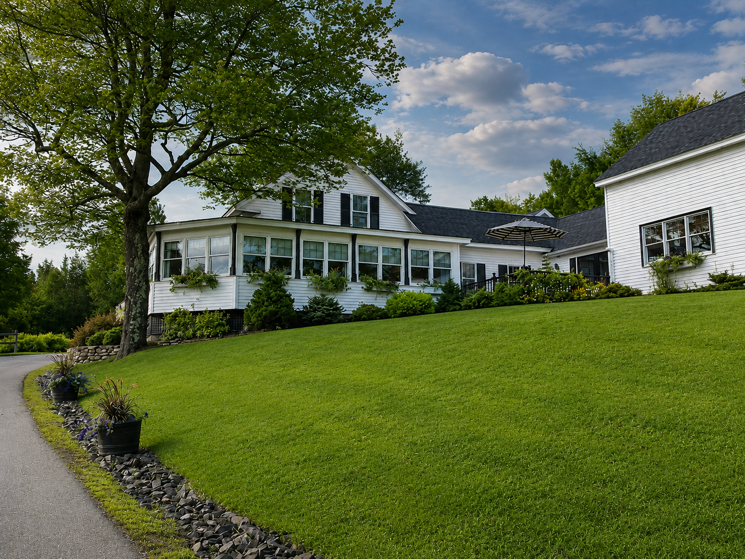 White house with porch and green lawn on a sloping hillside under a partly cloudy sky