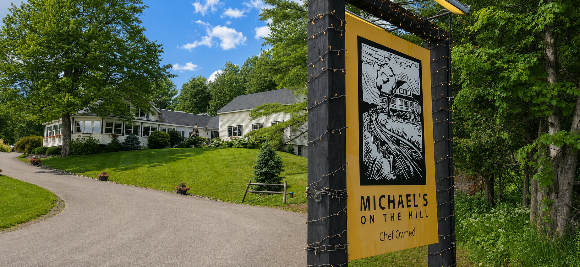 Entrance sign for Michael's on a wood driveway, with a white building and green lawn in the background