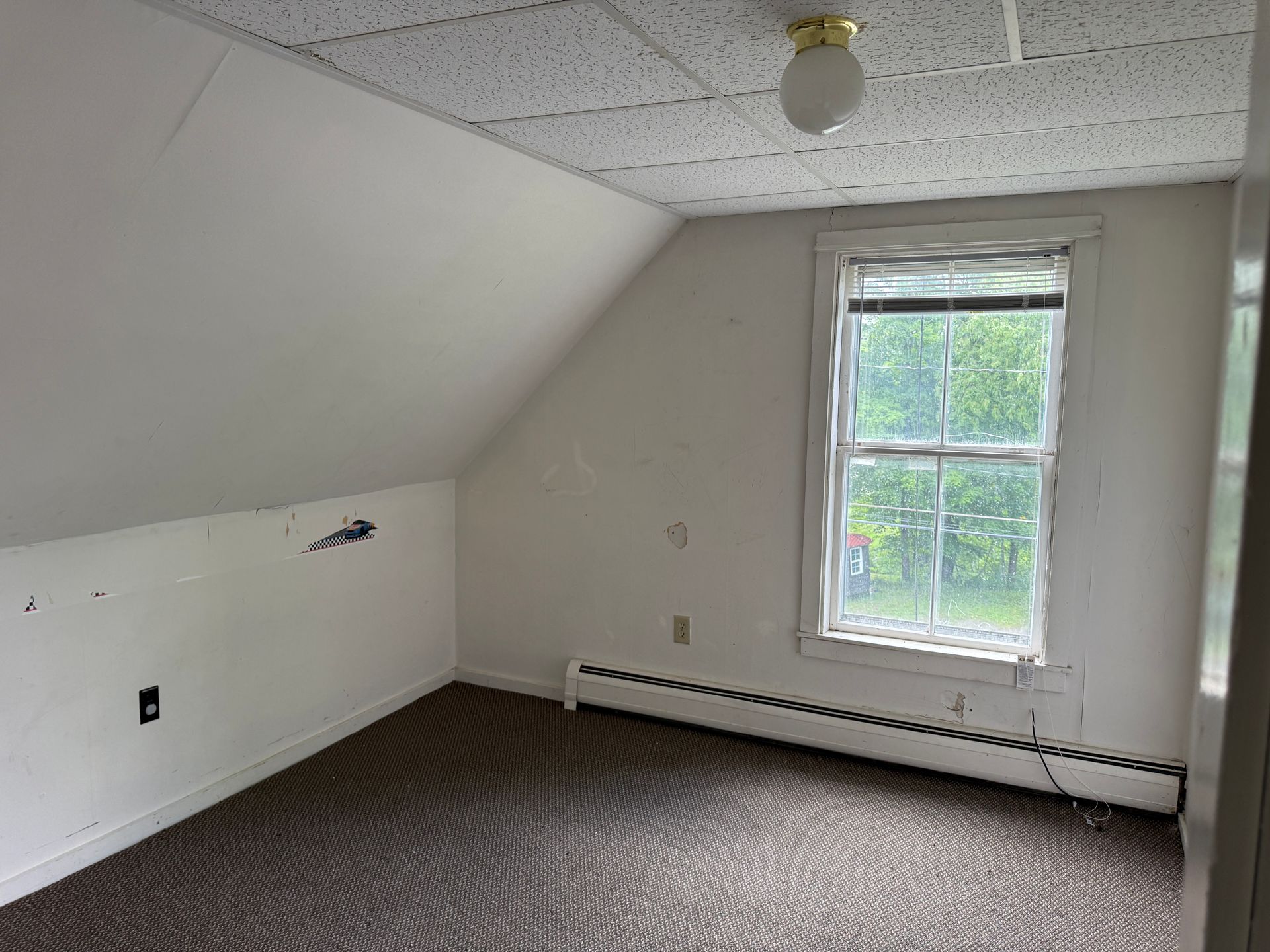 Empty attic bedroom with slanted white ceiling, small window, and gray carpet