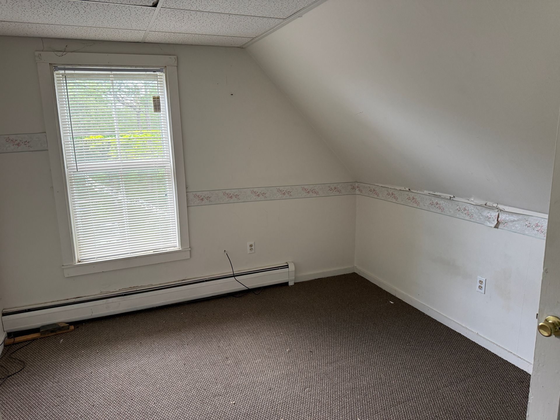 Empty attic room with slanted ceiling, window, white walls, and carpeted floor.