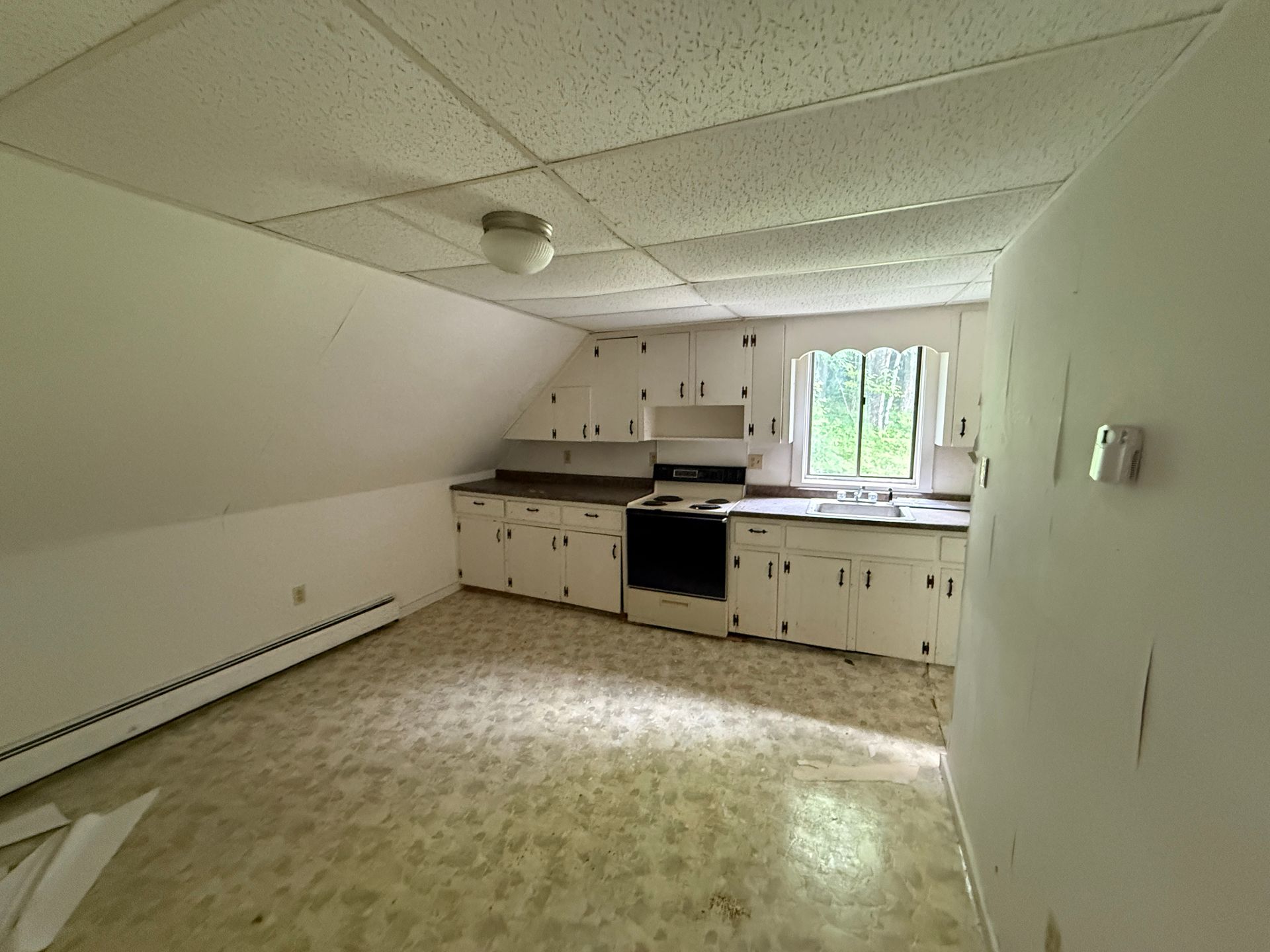 Empty kitchen with slanted ceiling, white cabinets, black stove, and window letting in daylight.