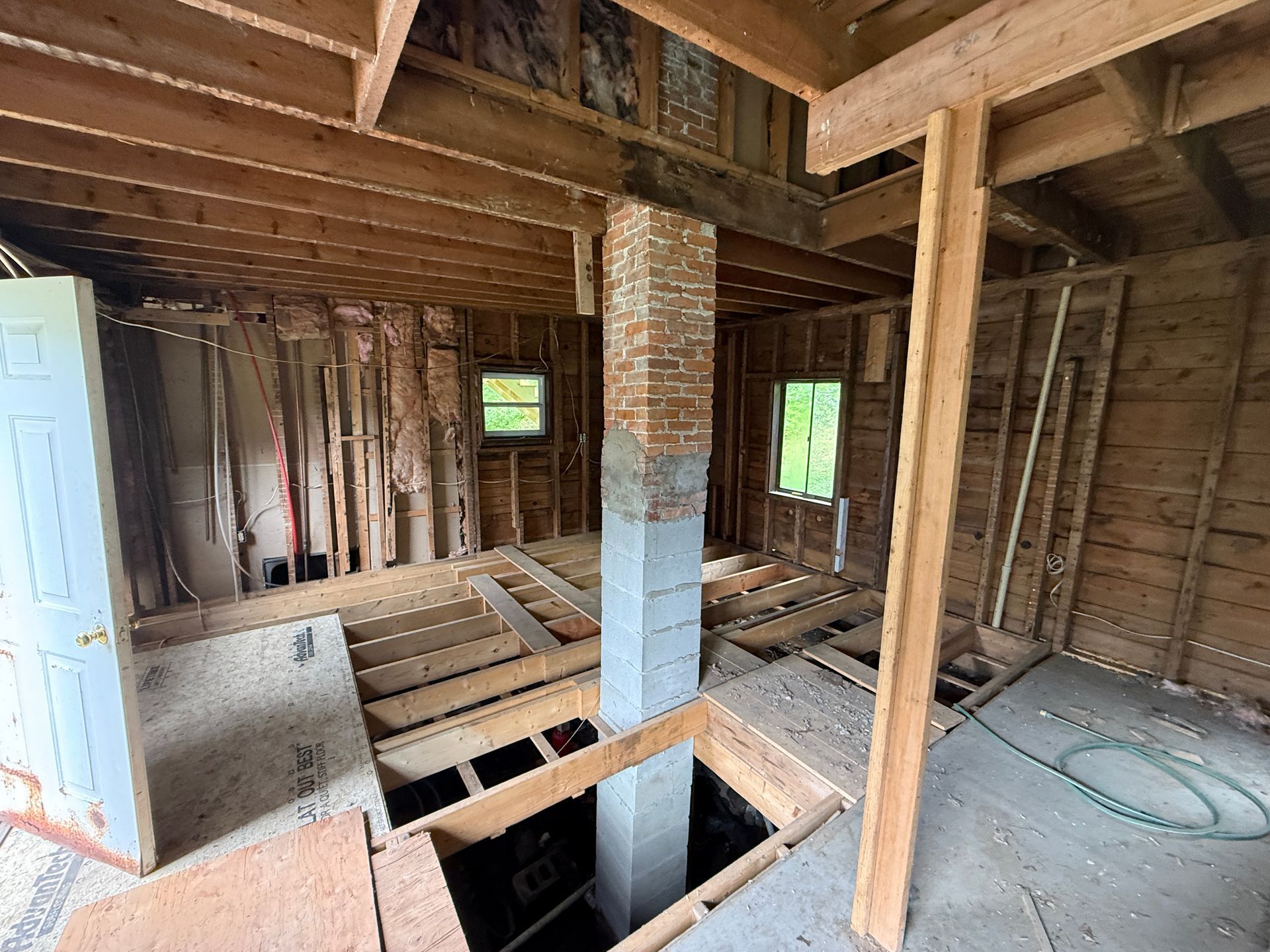 Unfinished house interior with exposed framing, brick chimney, open floor joists, and plywood subfloor.