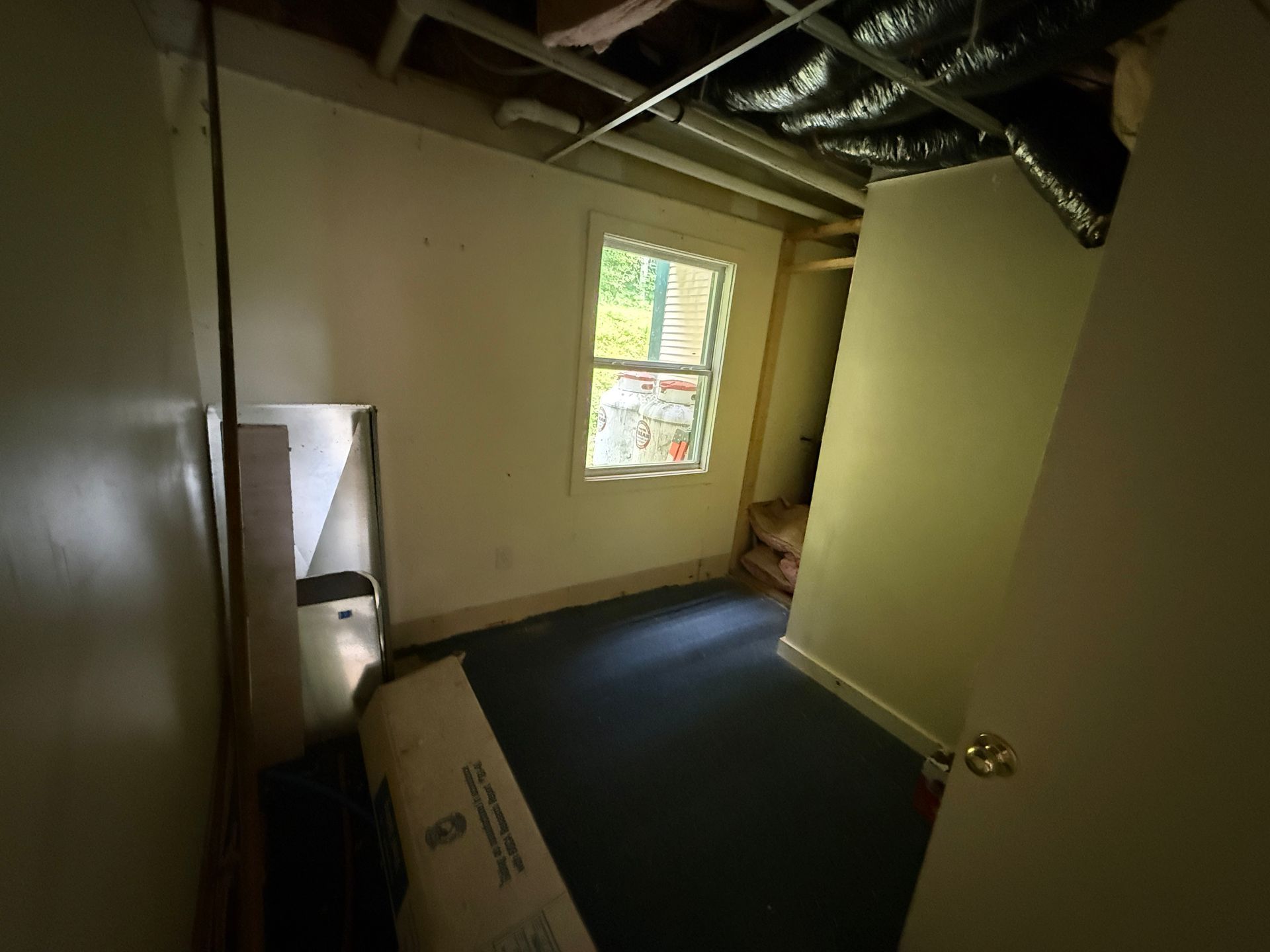 Empty basement room with bare walls, exposed pipes, and a small window letting in daylight.