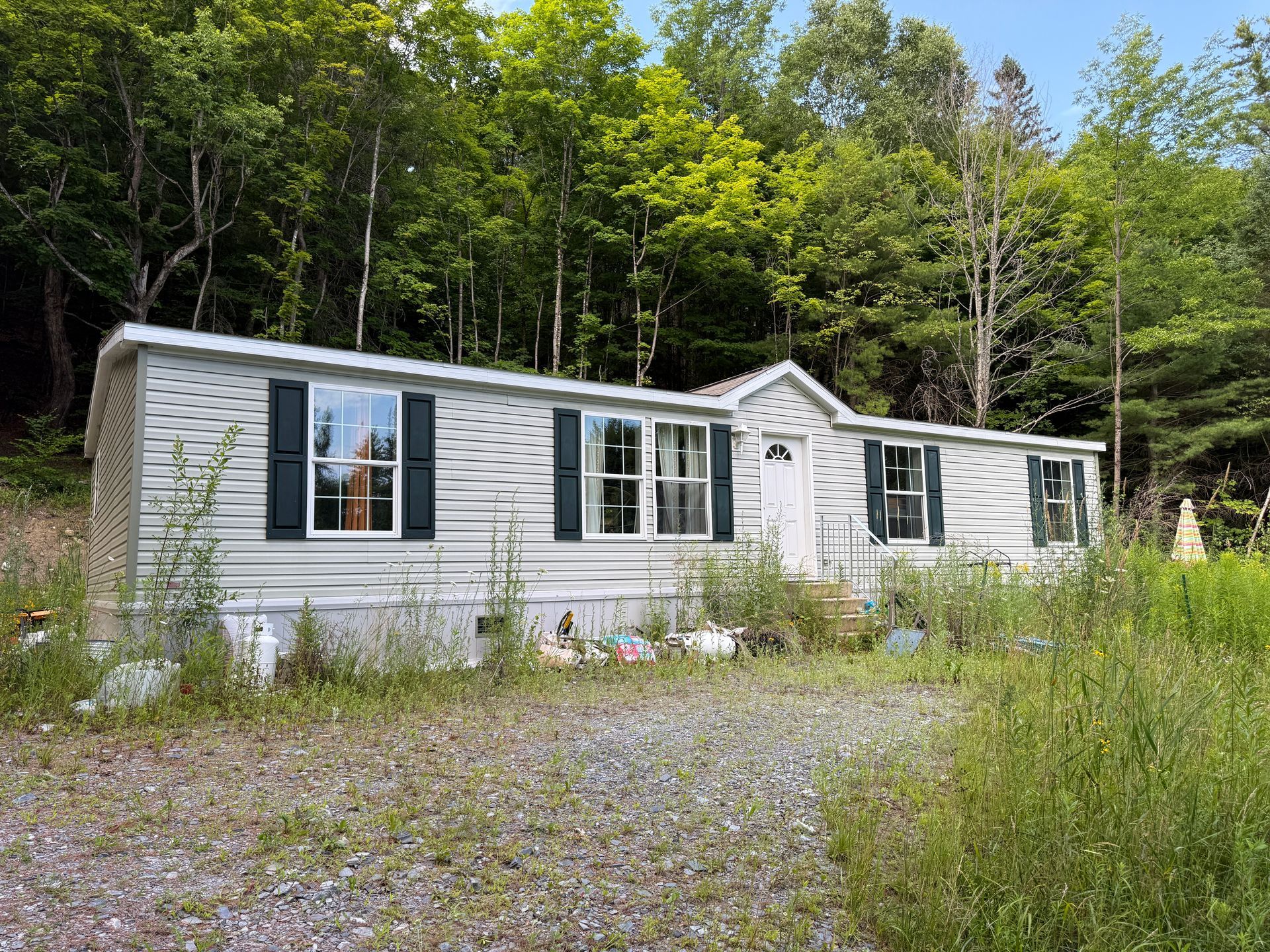White mobile home in a wooded clearing with a gravel driveway and tall grass.