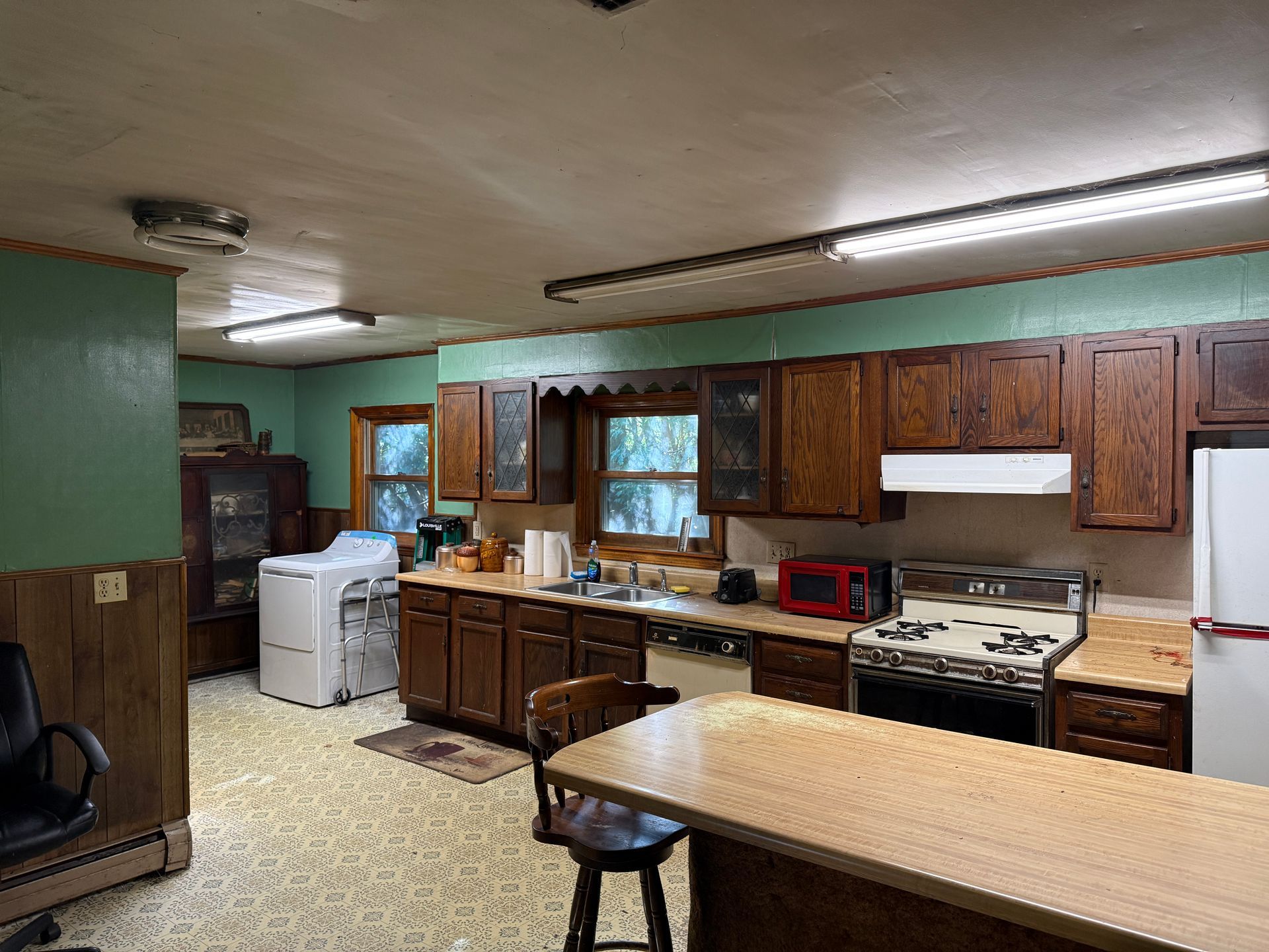 Modest kitchen with wood cabinets, white appliances, and a beige island under fluorescent lights.