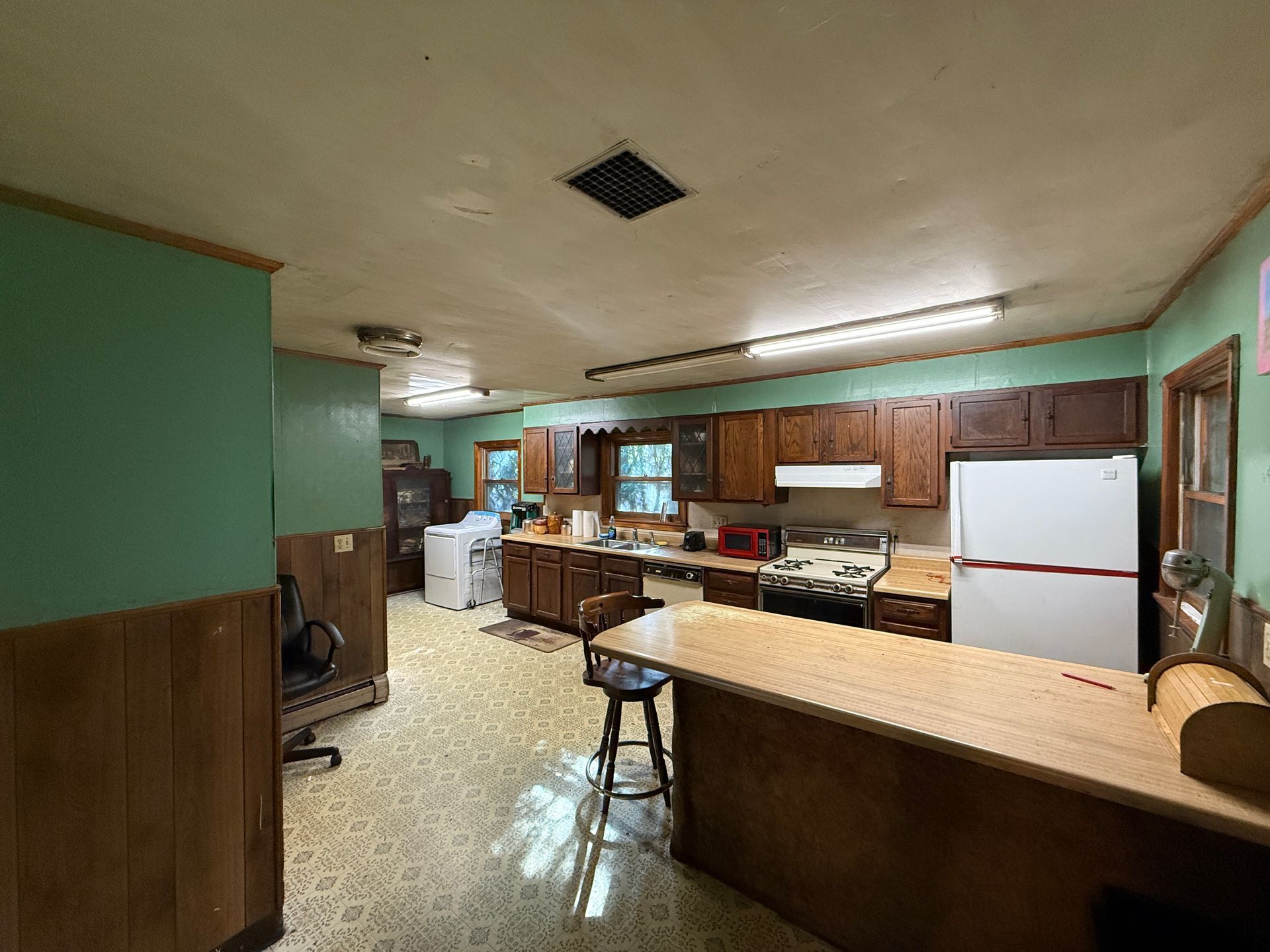 Wide kitchen with wood cabinets, white appliances, and a large island under fluorescent lights.