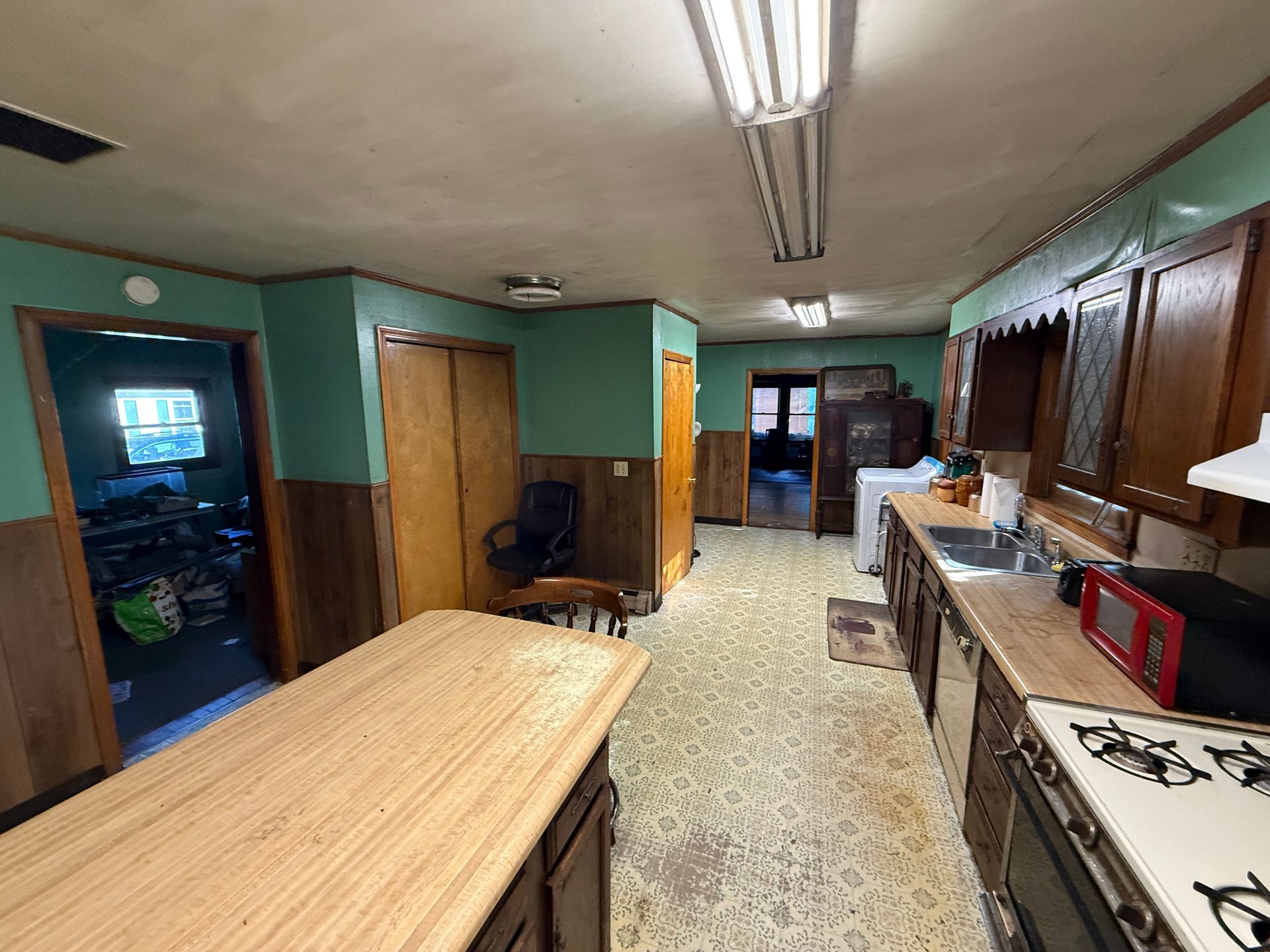 Long narrow kitchen with green walls, wooden cabinets, fluorescent lights, and a large butcher-block table