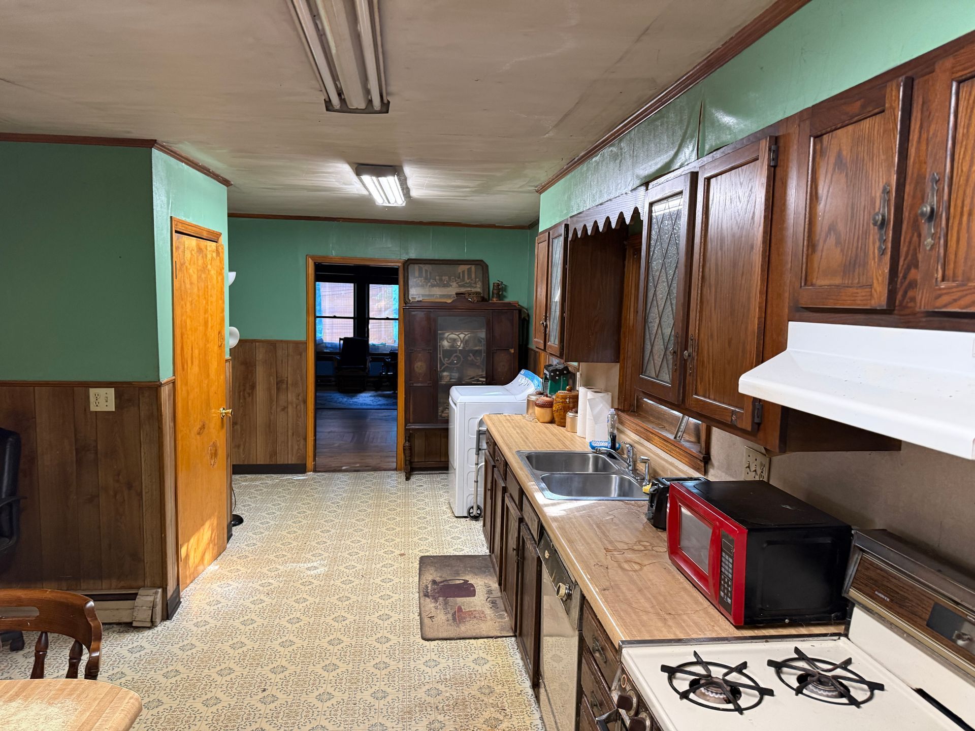 Narrow kitchen with wood cabinets, green walls, white stove, red microwave, and a small table at the far end