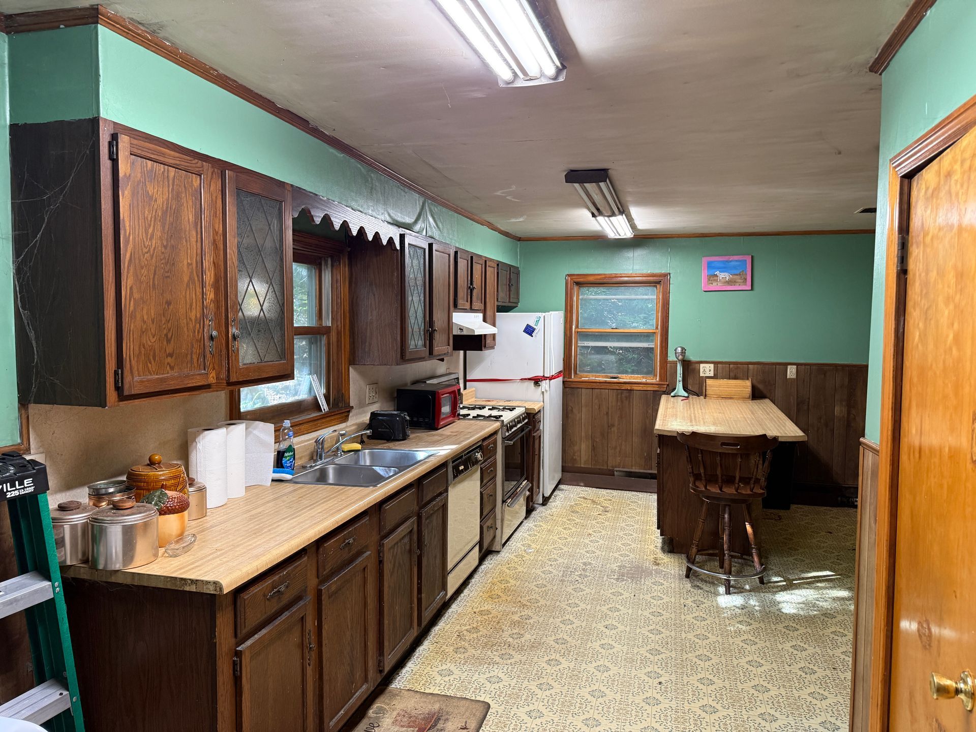 Long, narrow kitchen with wood cabinets, countertops, sink, stove, fridge, and a small table by the window.