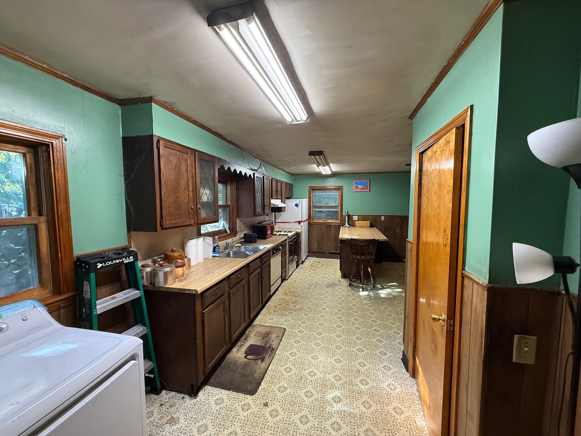 Narrow kitchen with mint-green walls, wood cabinets, fluorescent lights, and a dining table at the far end