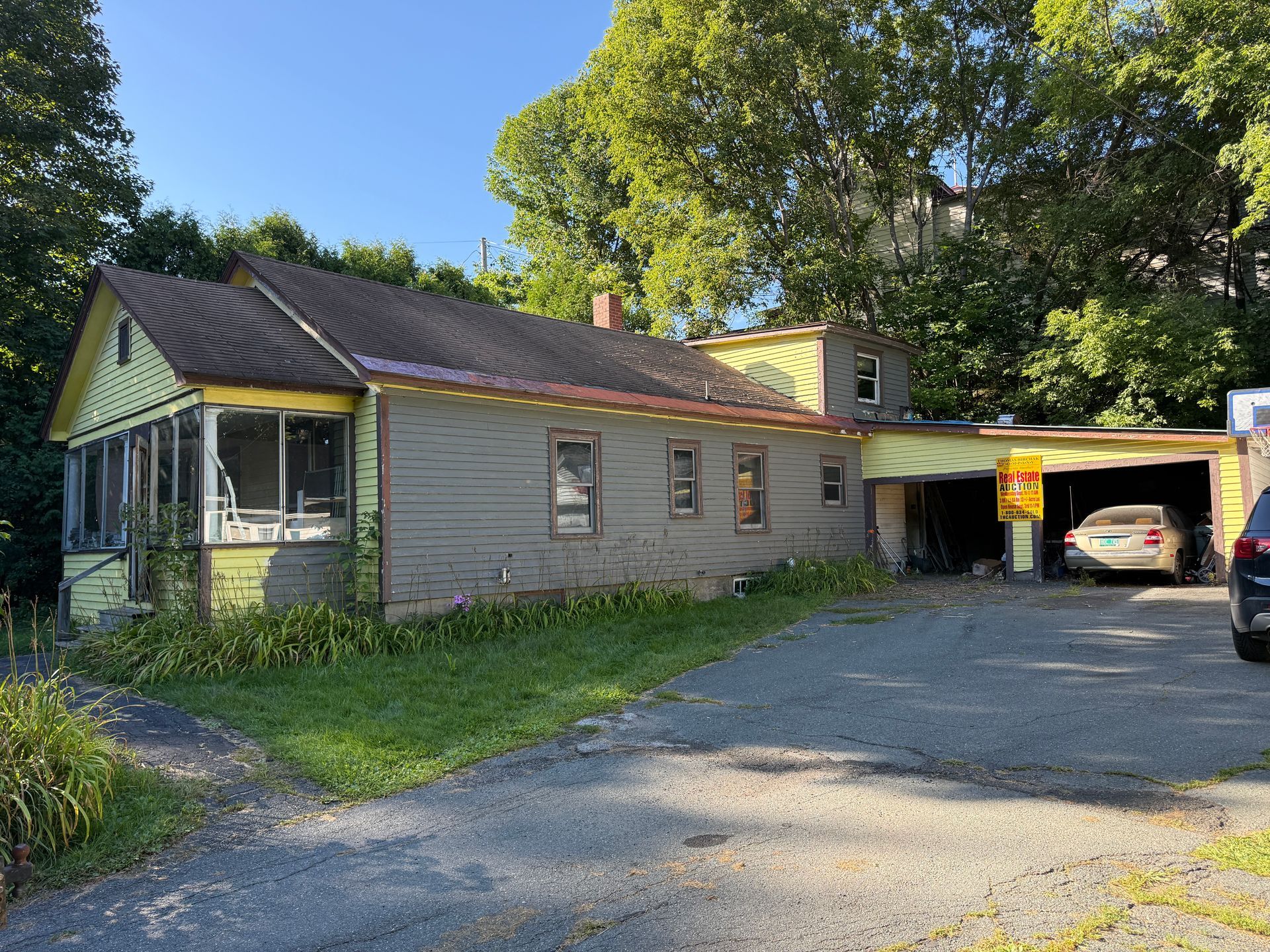 gray house with a carport, gravel driveway, and trees in the yard under a blue sky