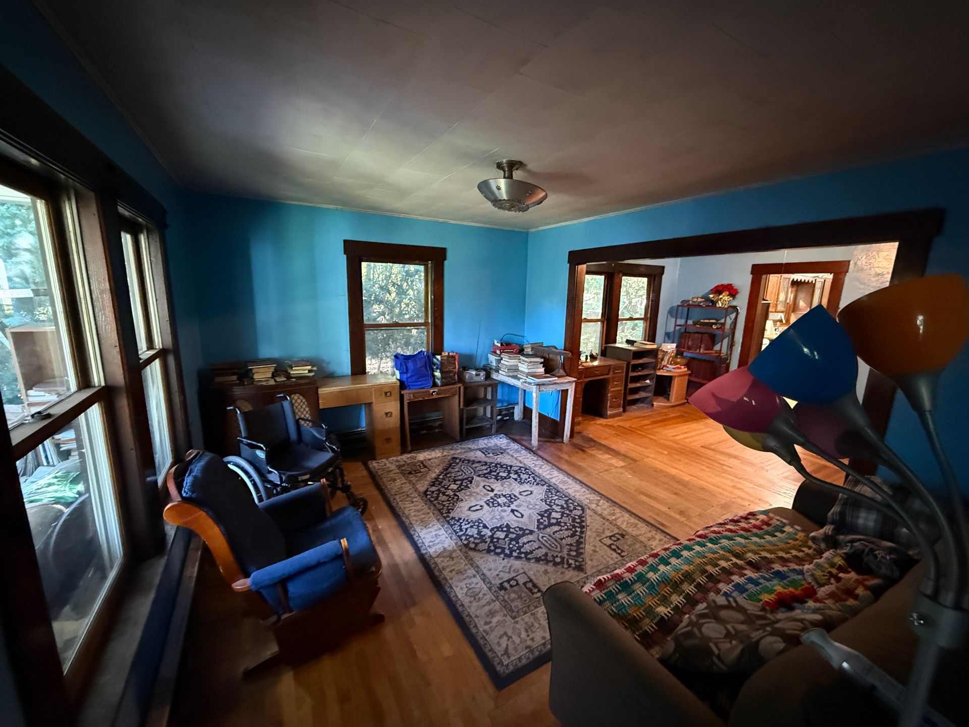 Cozy living room with blue walls, wood floors, rugs, chairs, desks, and a floor lamp by a large window.