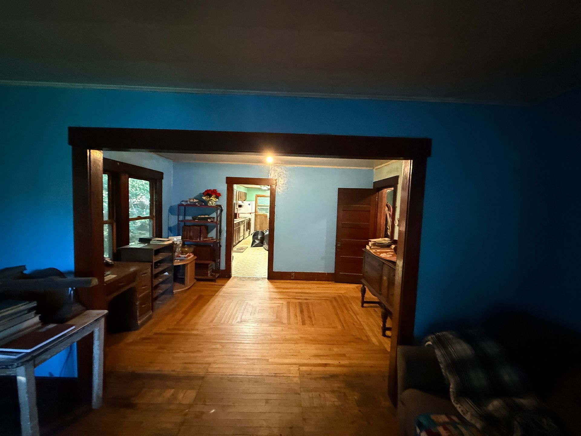 Blue-walled living room with wooden floor, doorway to a lit kitchen, and furniture along the sides
