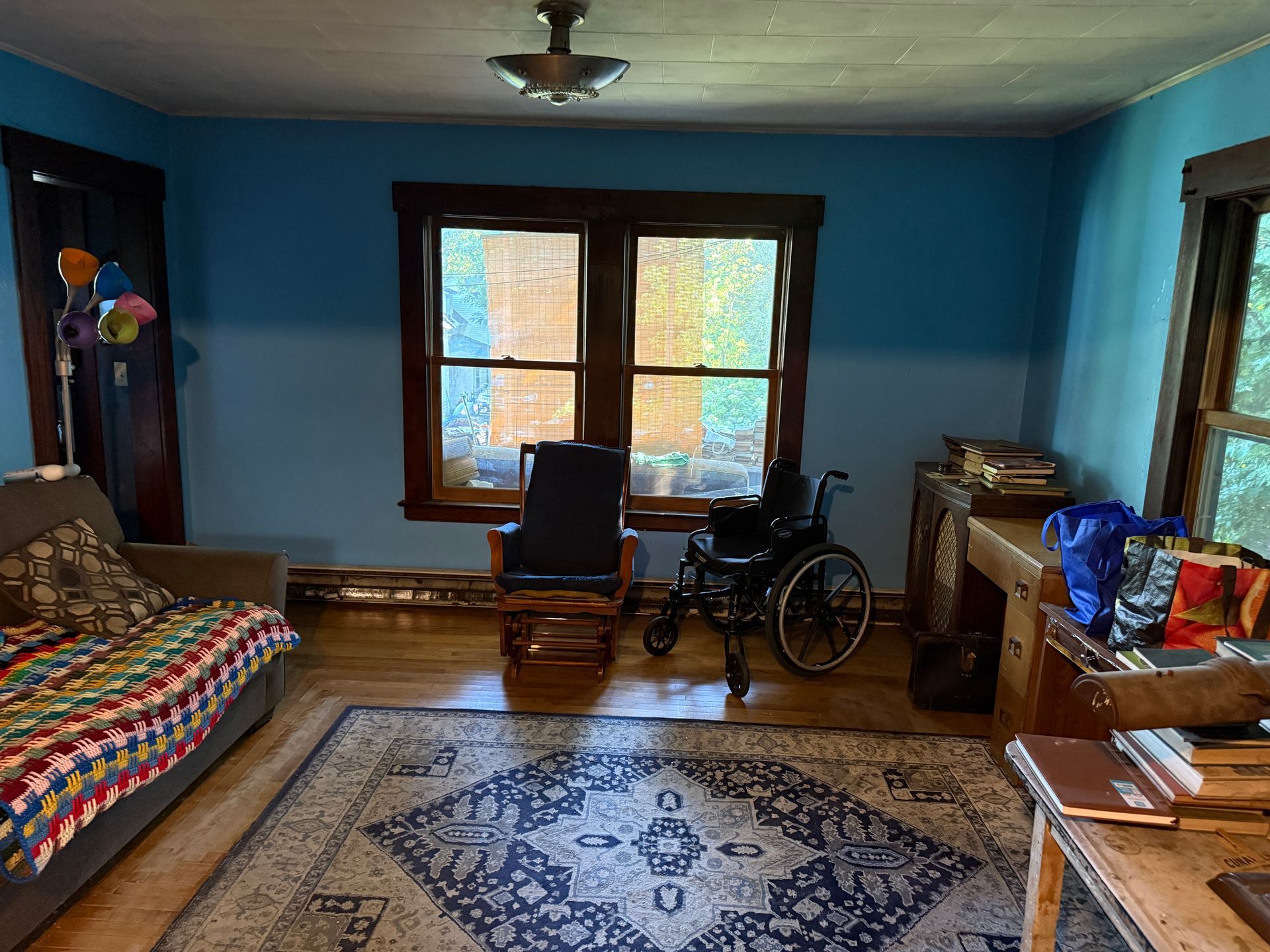 Blue room with windows, rug, chair, wheelchair, and a patterned bedspread.