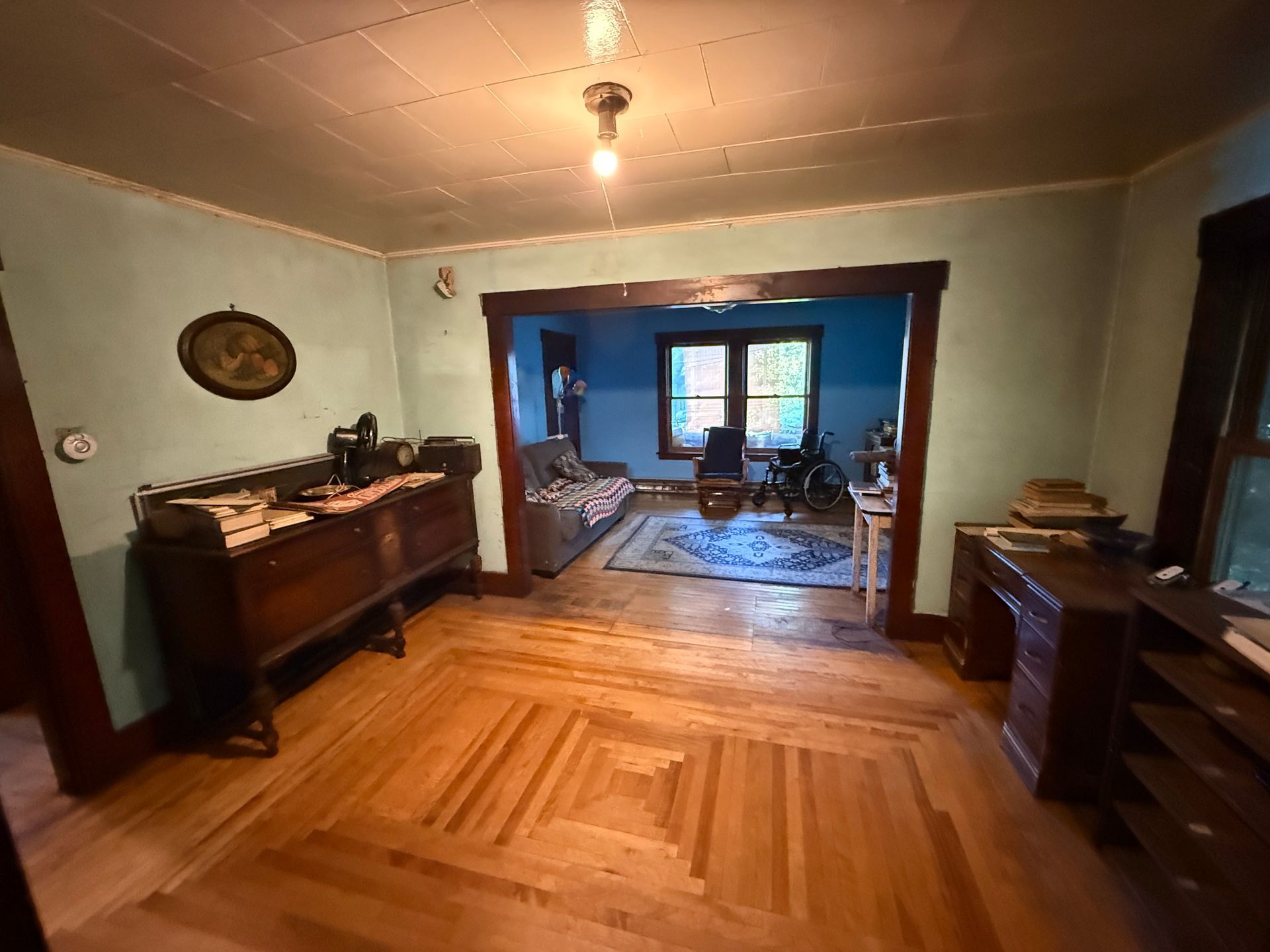 Old room with wooden floor, vintage furniture, and a doorway to a blue sitting room with windows.