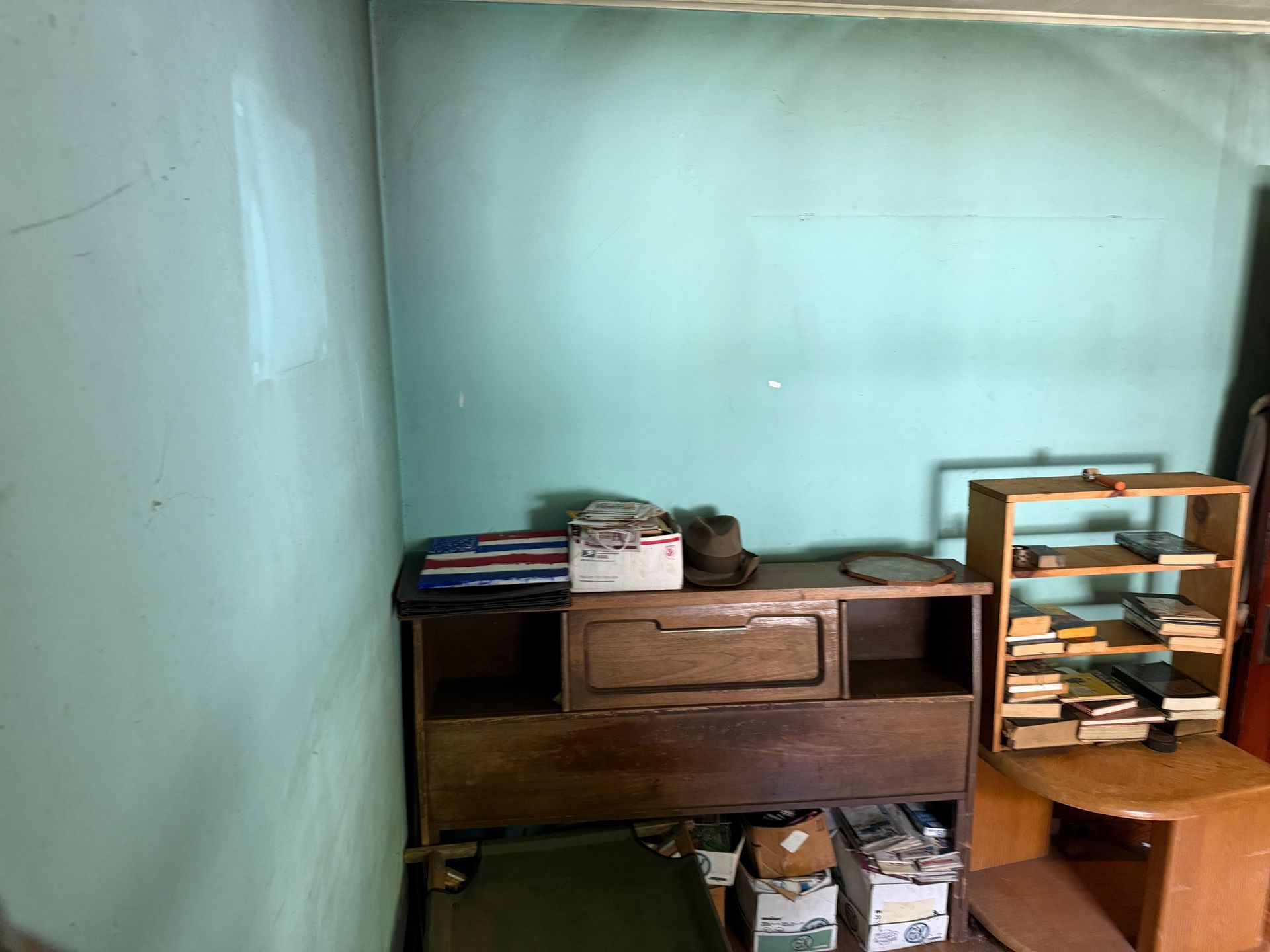 Small, sparse room with a wooden dresser, stacked books, and a shelf against pale green walls.