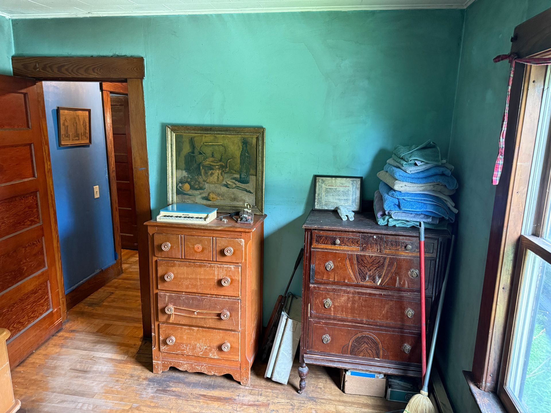 Small teal bedroom with wooden dressers, a framed painting, and bags stacked by the wall.