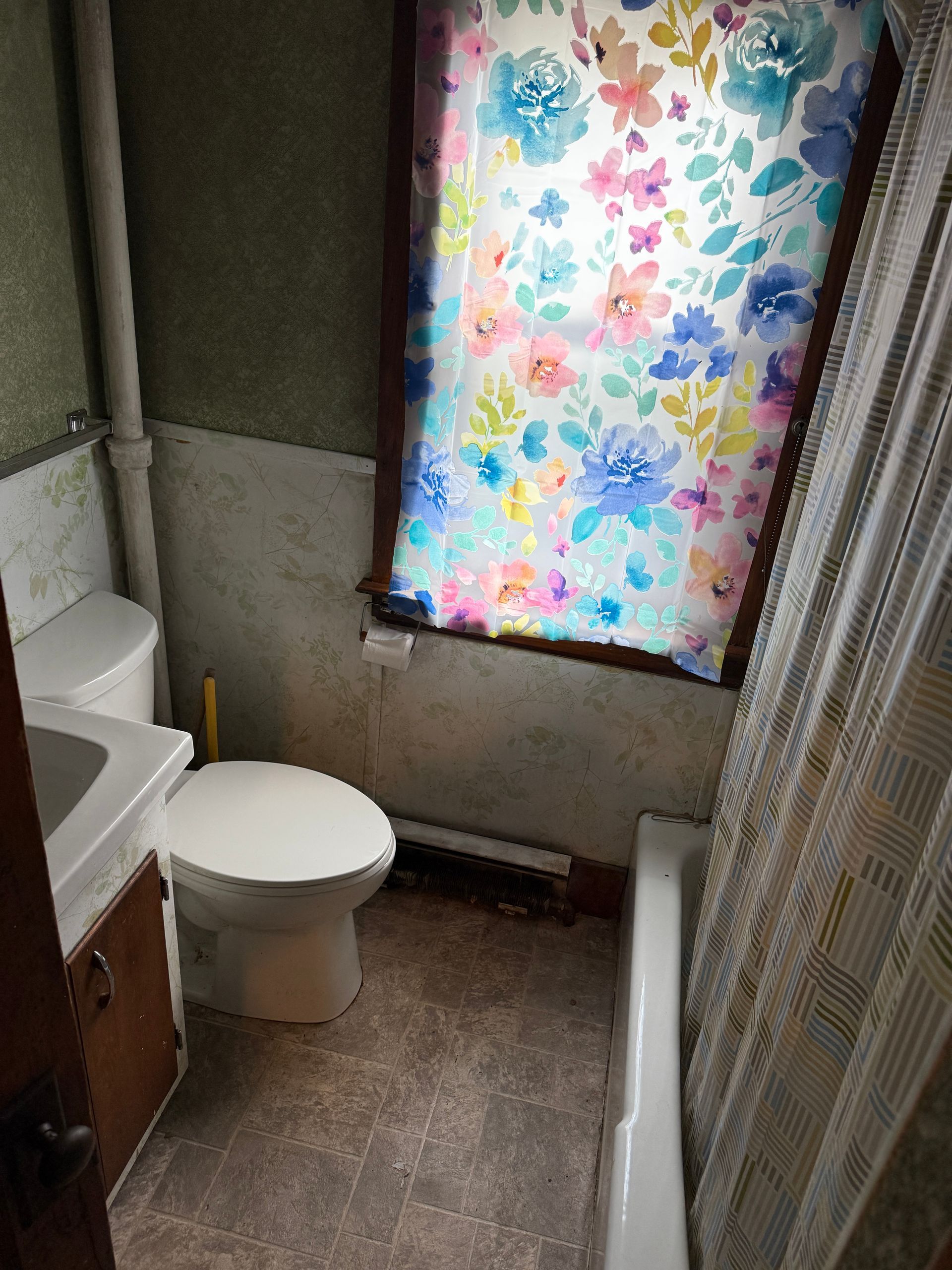 Small bathroom with toilet, sink, shower curtain, and colorful floral window covering.
