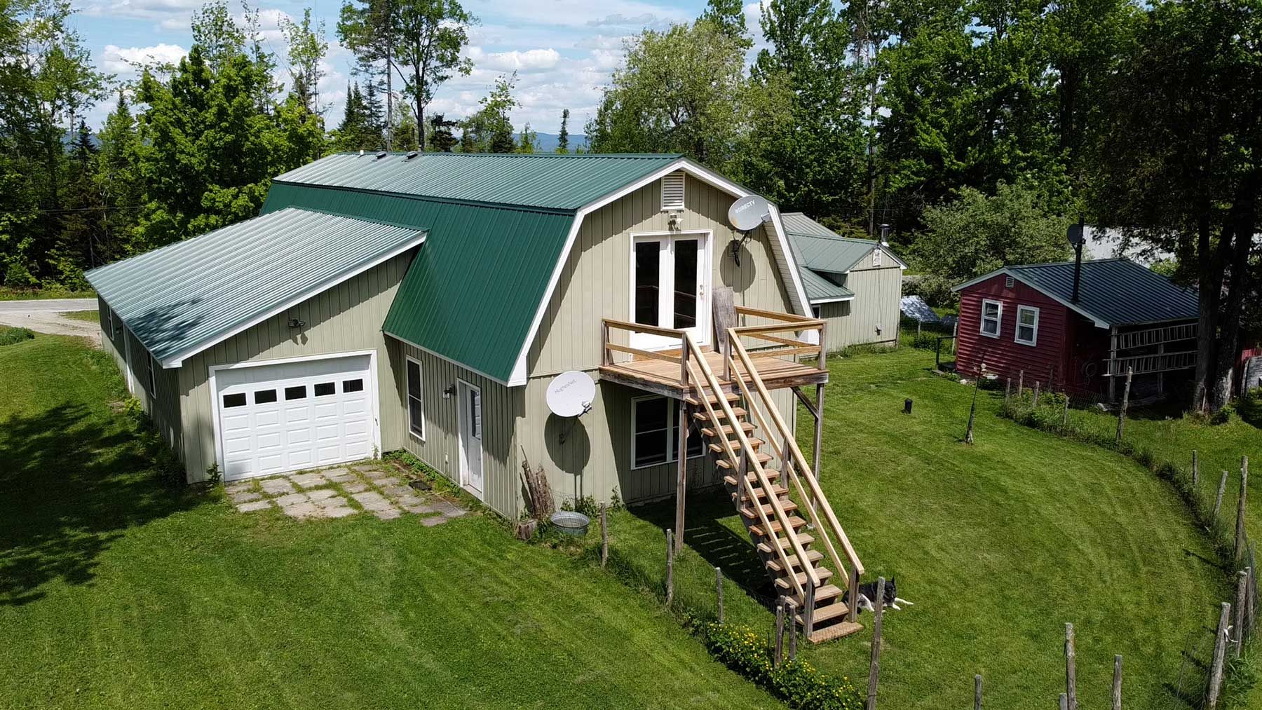 Green-roofed barn-style house with garage, wooden stairs, and small red building on a grassy hill.