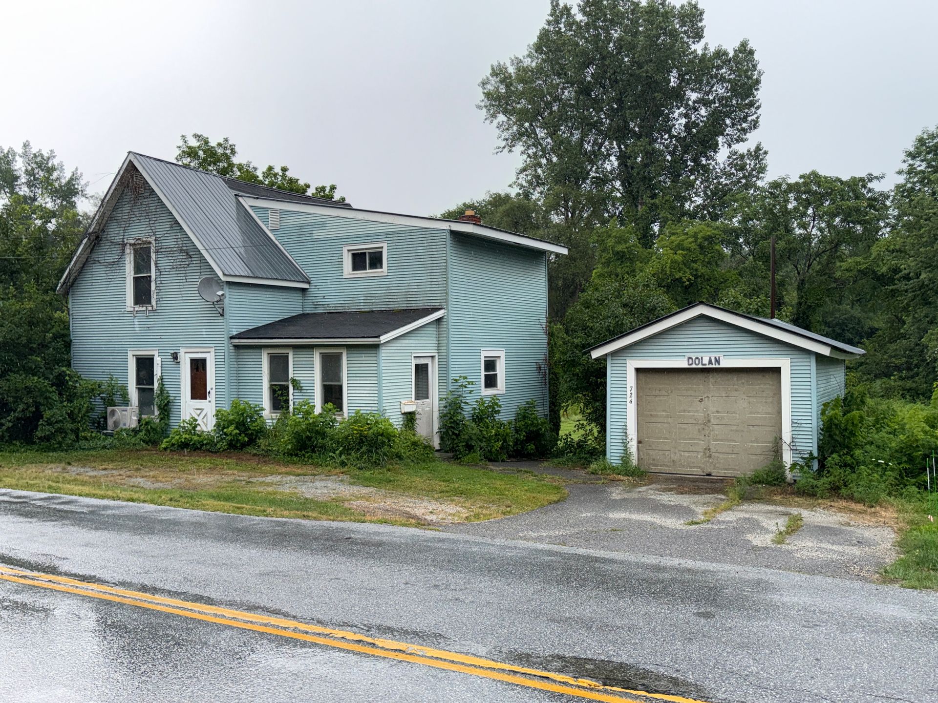 Blue two-story house with detached garage beside a wet road, surrounded by trees