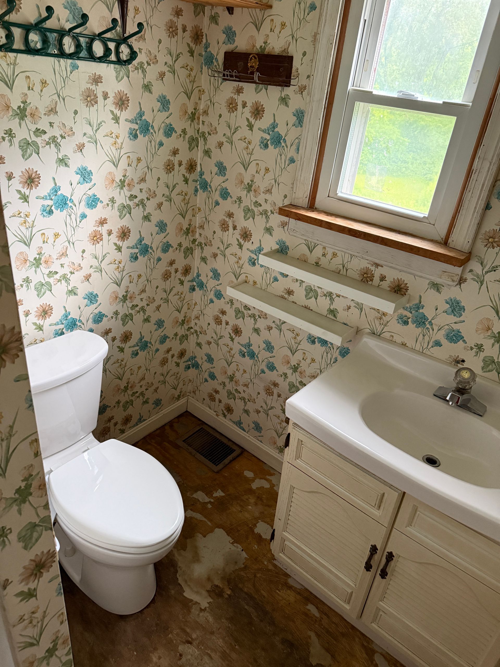 Small bathroom with floral wallpaper, toilet, sink vanity, window, and wall shelves.