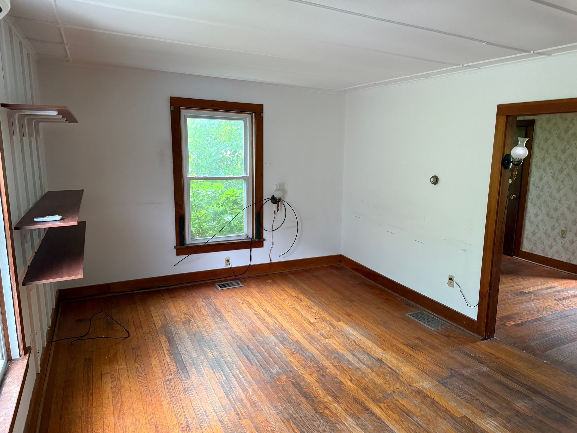 Empty room with hardwood floors, white walls, a window, and a doorway to an adjoining room.