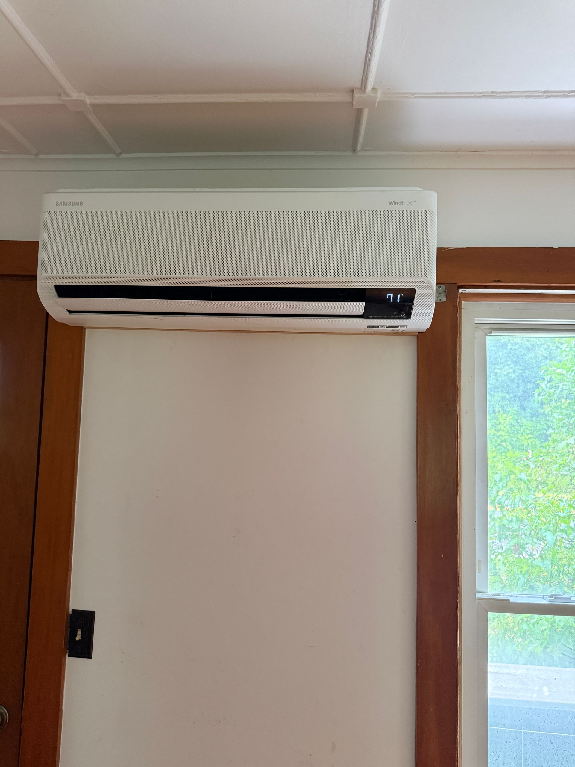 Wall-mounted air conditioner above a door in a beige room with wood trim and a window.