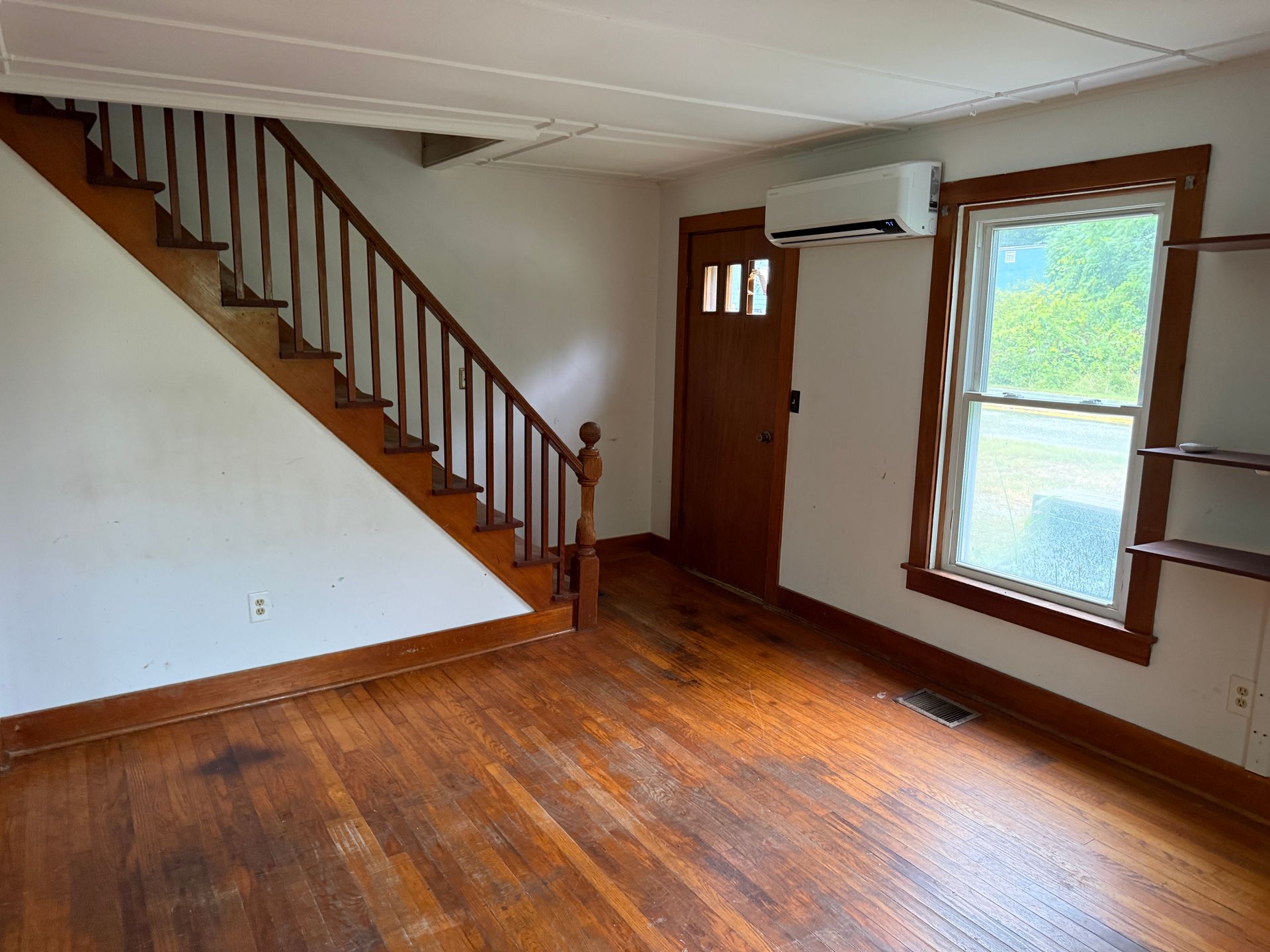 Empty living room with wood floors, staircase, front door, and window air conditioner