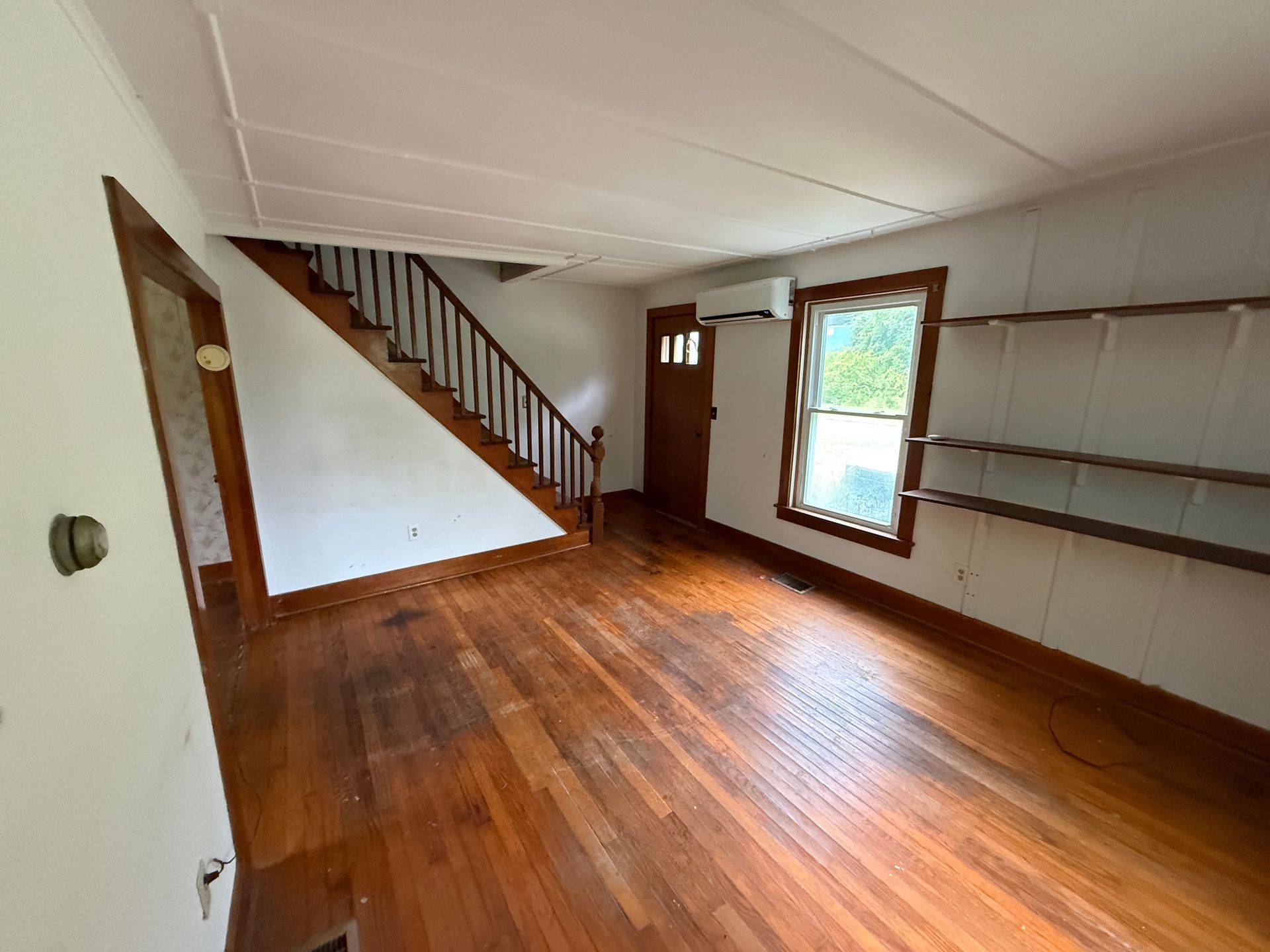 Empty room with hardwood floor, staircase, built-in shelves, and a window with daylight.