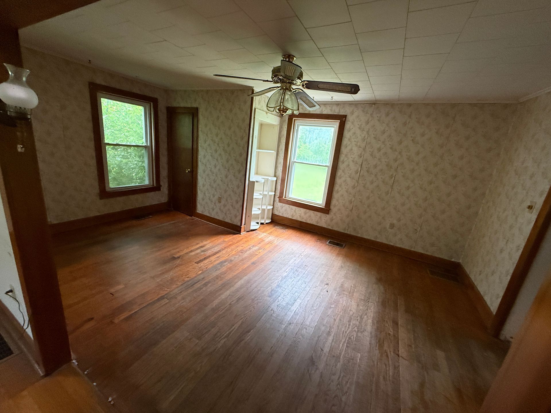 Empty room with hardwood floor, two windows, patterned wallpaper, and a ceiling fan with lights