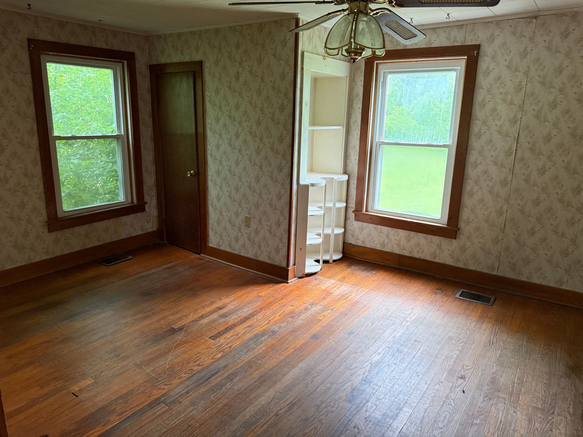 Empty room with hardwood floors, two windows, wallpaper, and a ceiling fan.