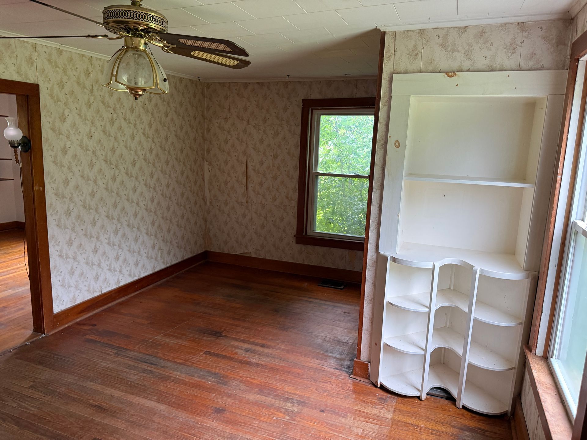 Empty room with worn wood floors, patterned wallpaper, a ceiling fan, and a built-in shelf by the window