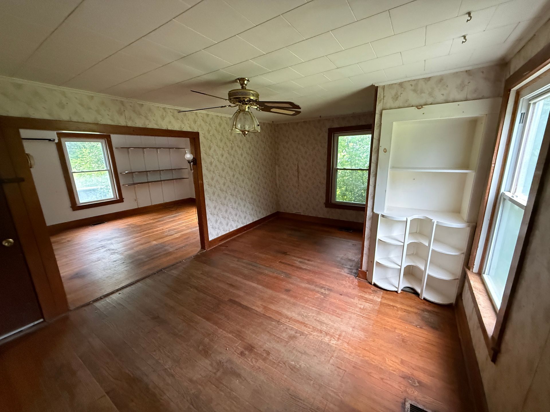 Empty wood-floored room with beige wallpaper, ceiling fan, windows, and built-in white shelves