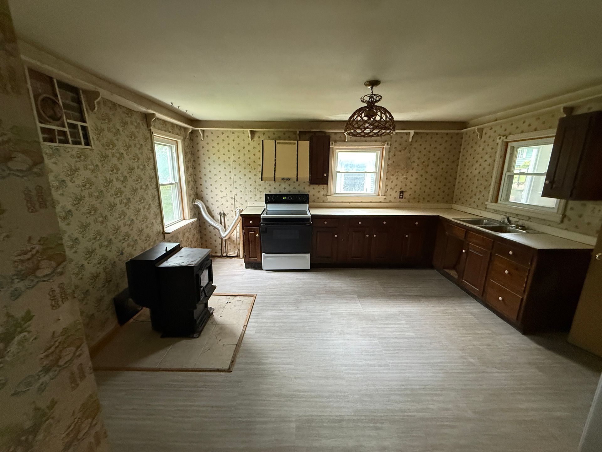 Empty vintage kitchen with wood cabinets, patterned wallpaper, and a black stove on the left