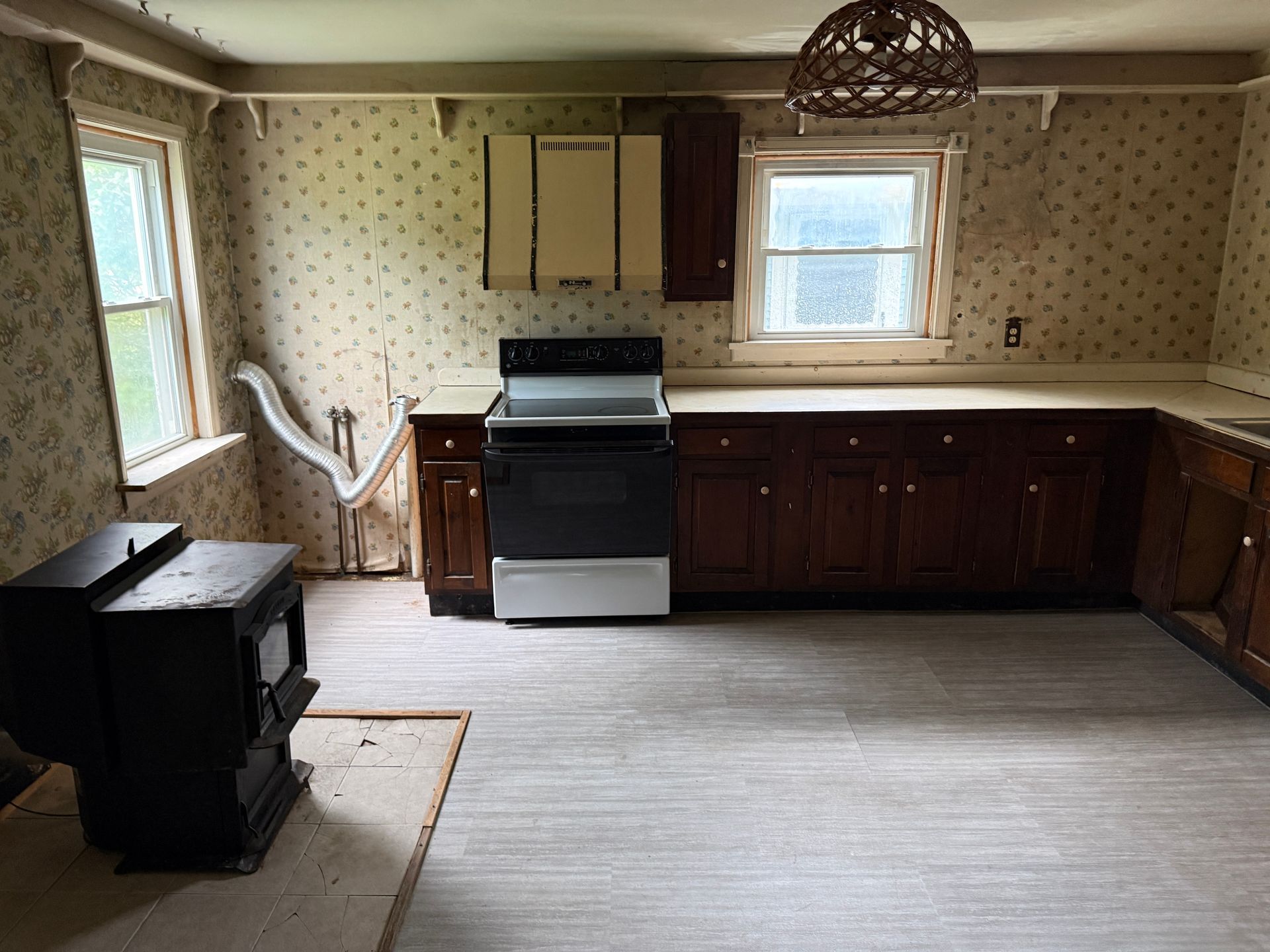 Empty vintage kitchen with brown cabinets, white stove, two windows, and a black wood stove on the left.