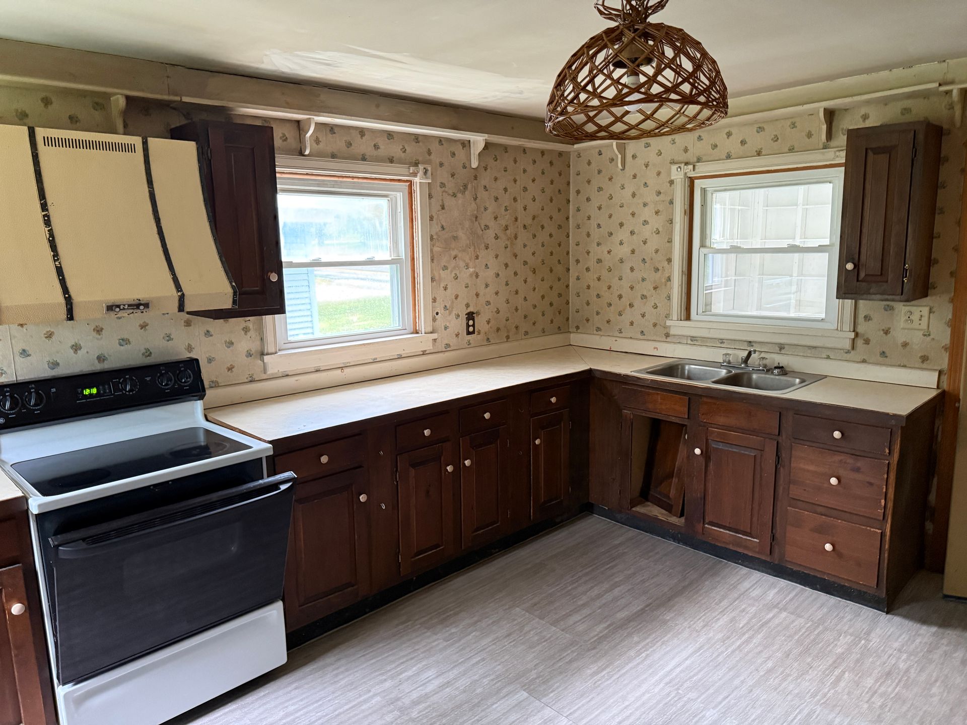 Empty kitchen with dark cabinets, white countertops, black stove, and two windows with patterned wallpaper.