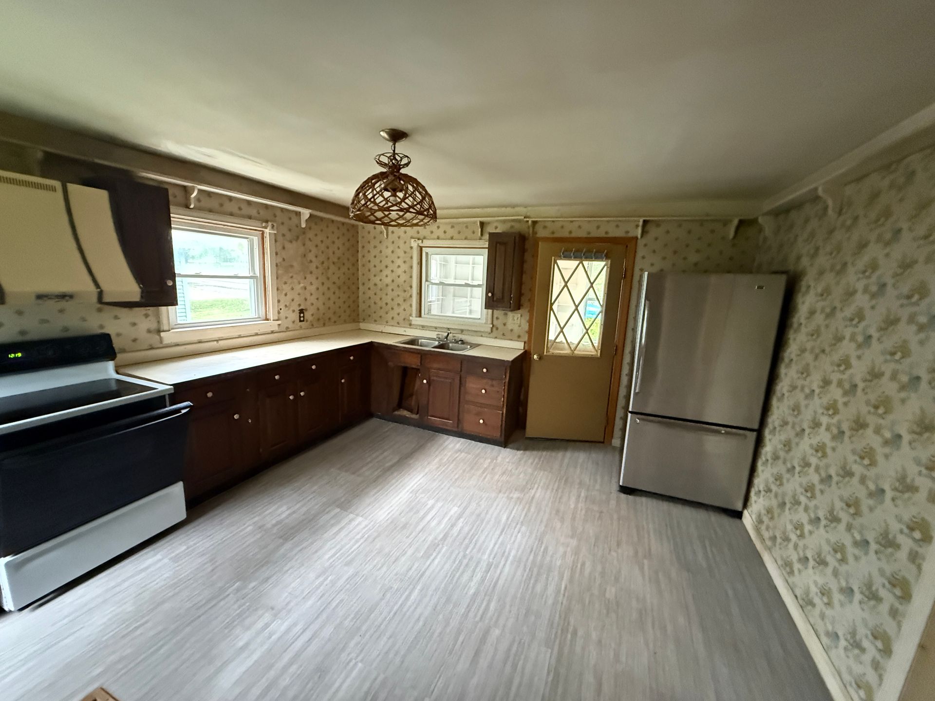 Empty kitchen with dark cabinets, white appliances, patterned wallpaper, and a door leading outside.