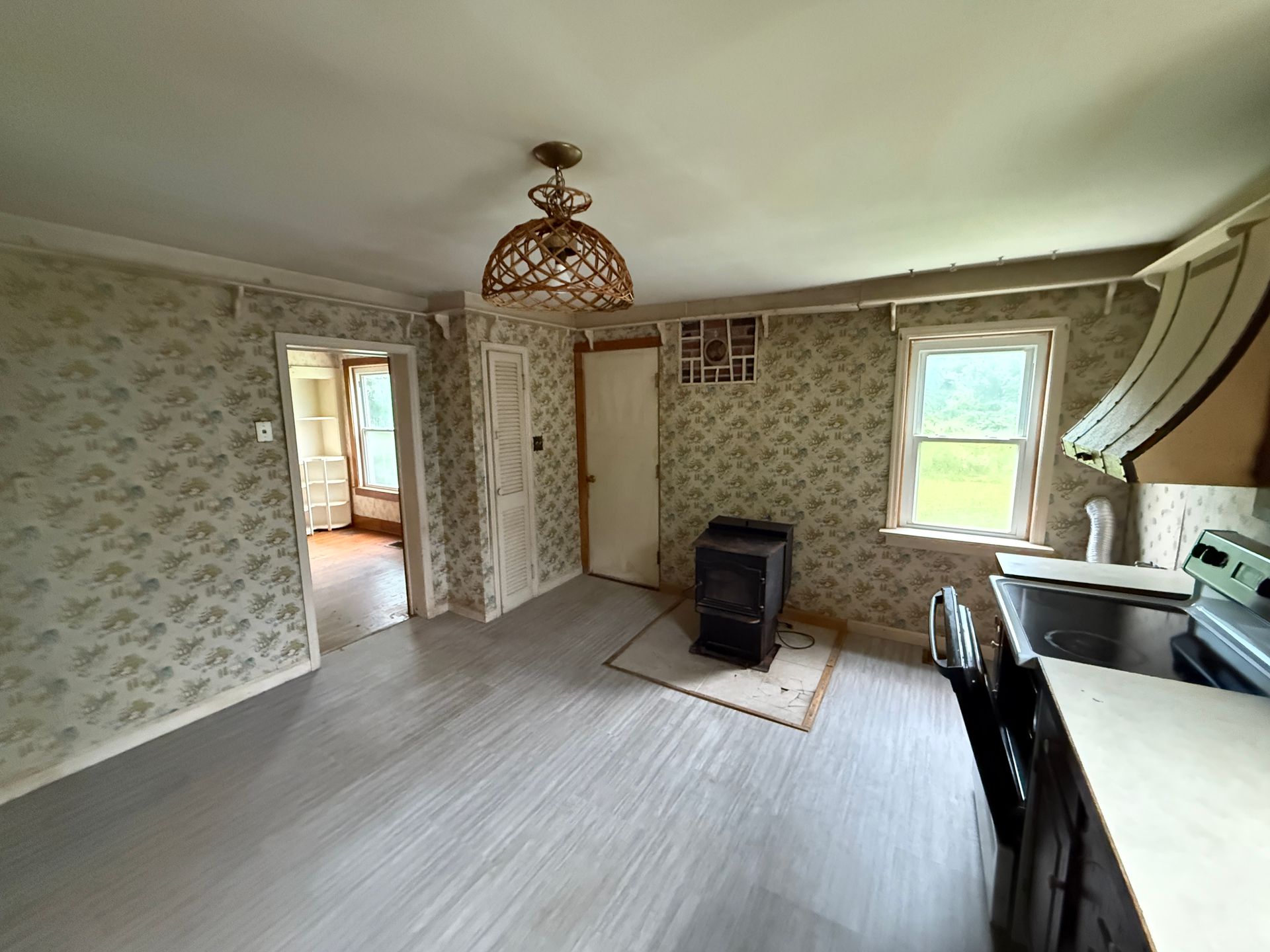 Empty room with floral wallpaper, two windows, a hanging light fixture, and a small stove on the floor.