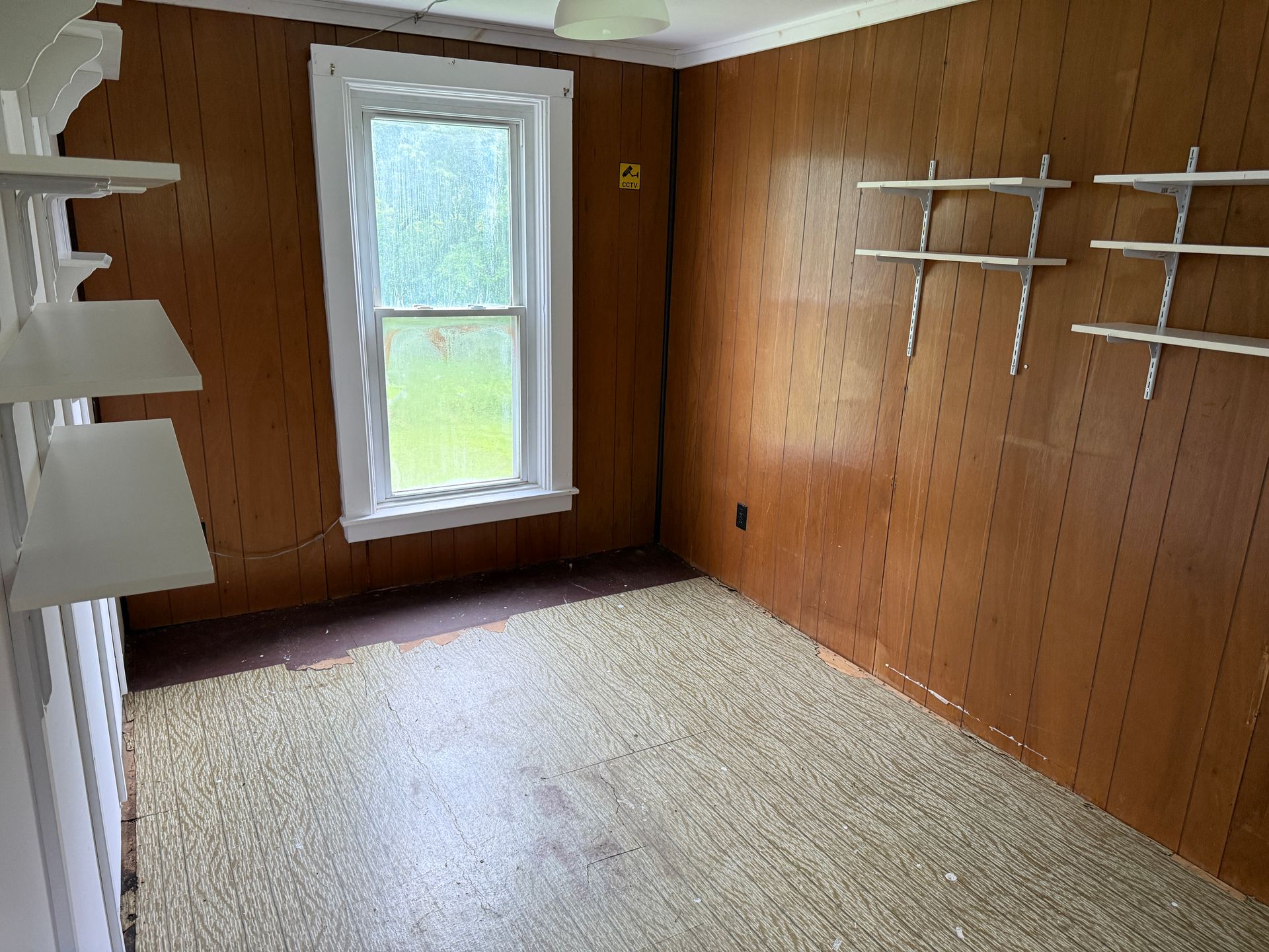 Empty small room with wood-paneled walls, beige carpet, window, and white shelves on the left and right.