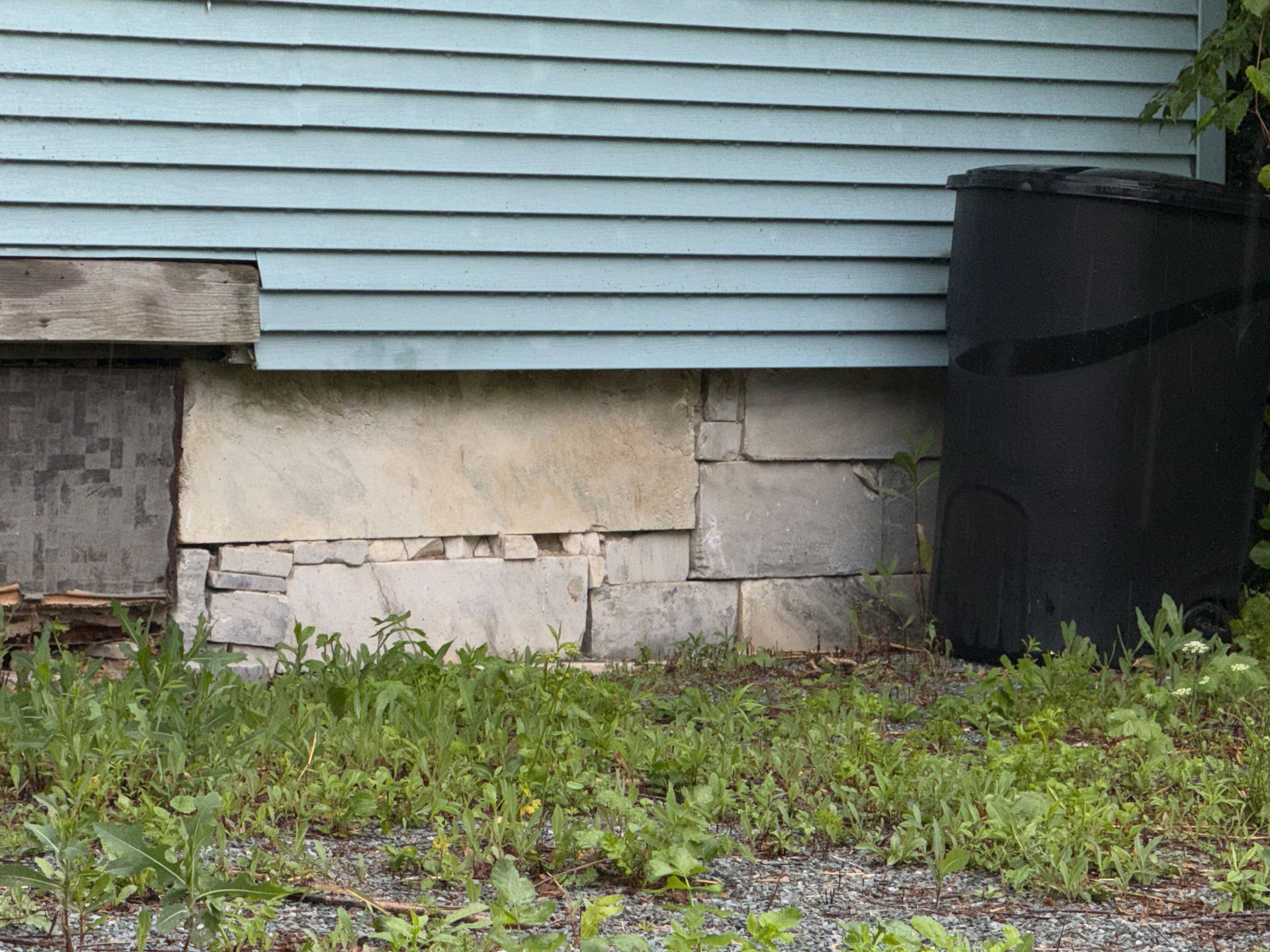 House foundation with stone and concrete blocks, blue siding above, and a black rain barrel on the right.