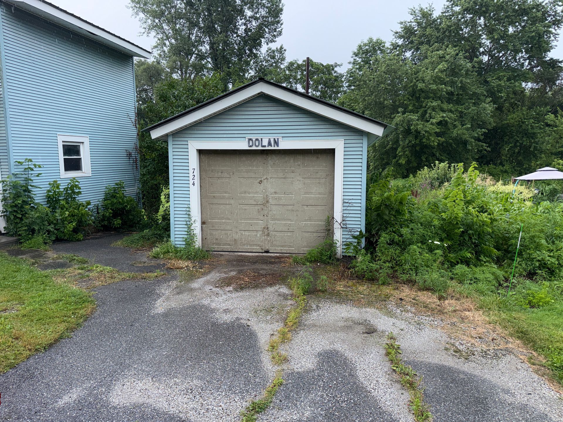 Small white garage with a closed beige door beside a driveway and greenery.