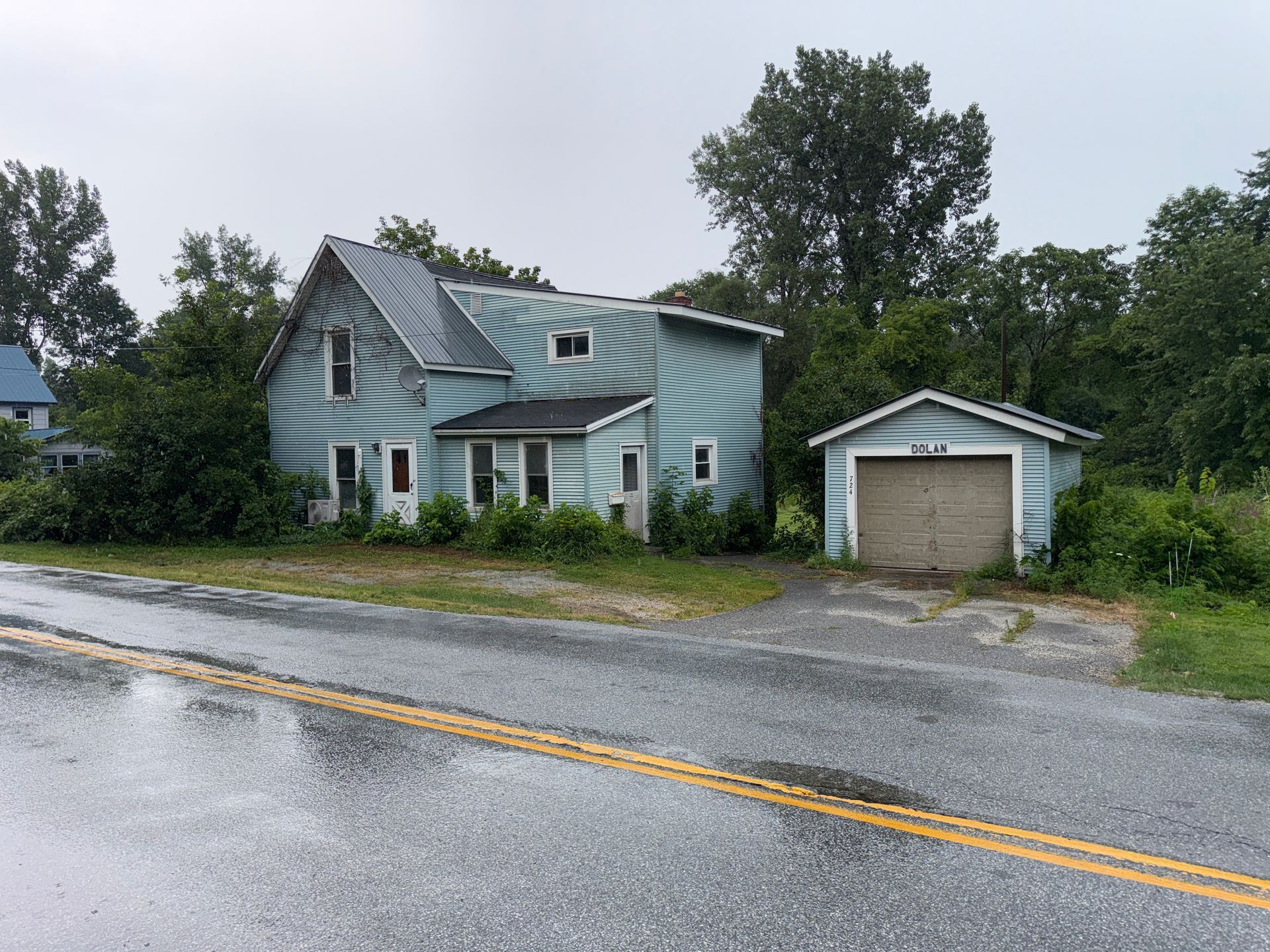 Rainy roadside view of a light blue house and detached garage with wet pavement and trees behind them