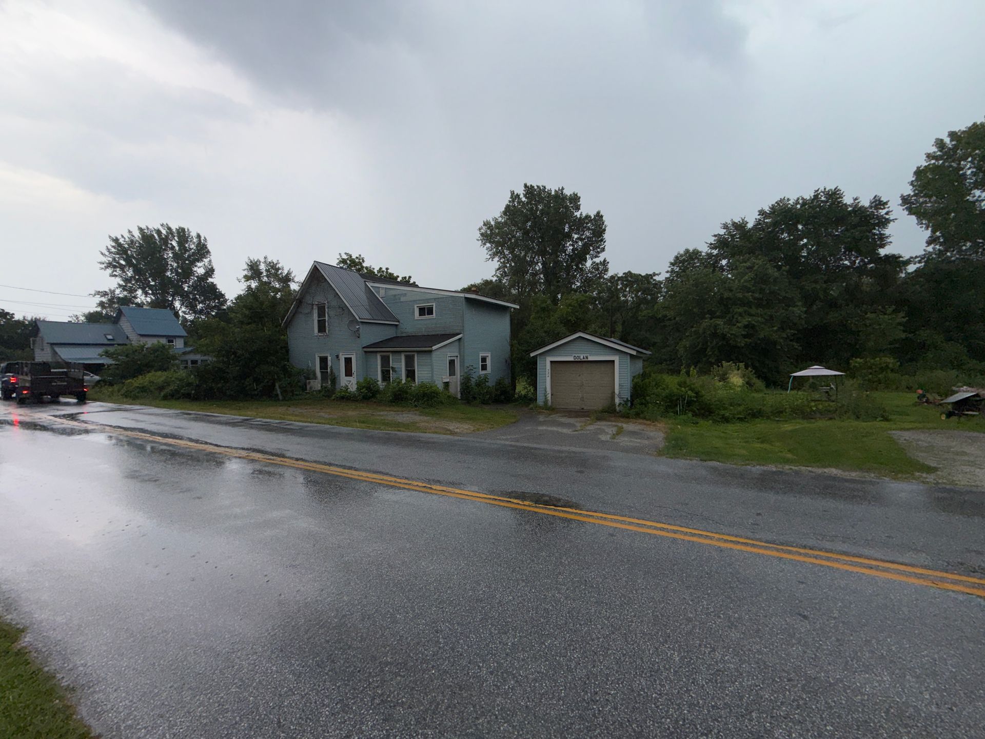 Rainy street with a small gray house, detached garage, and trees beside a wet road.