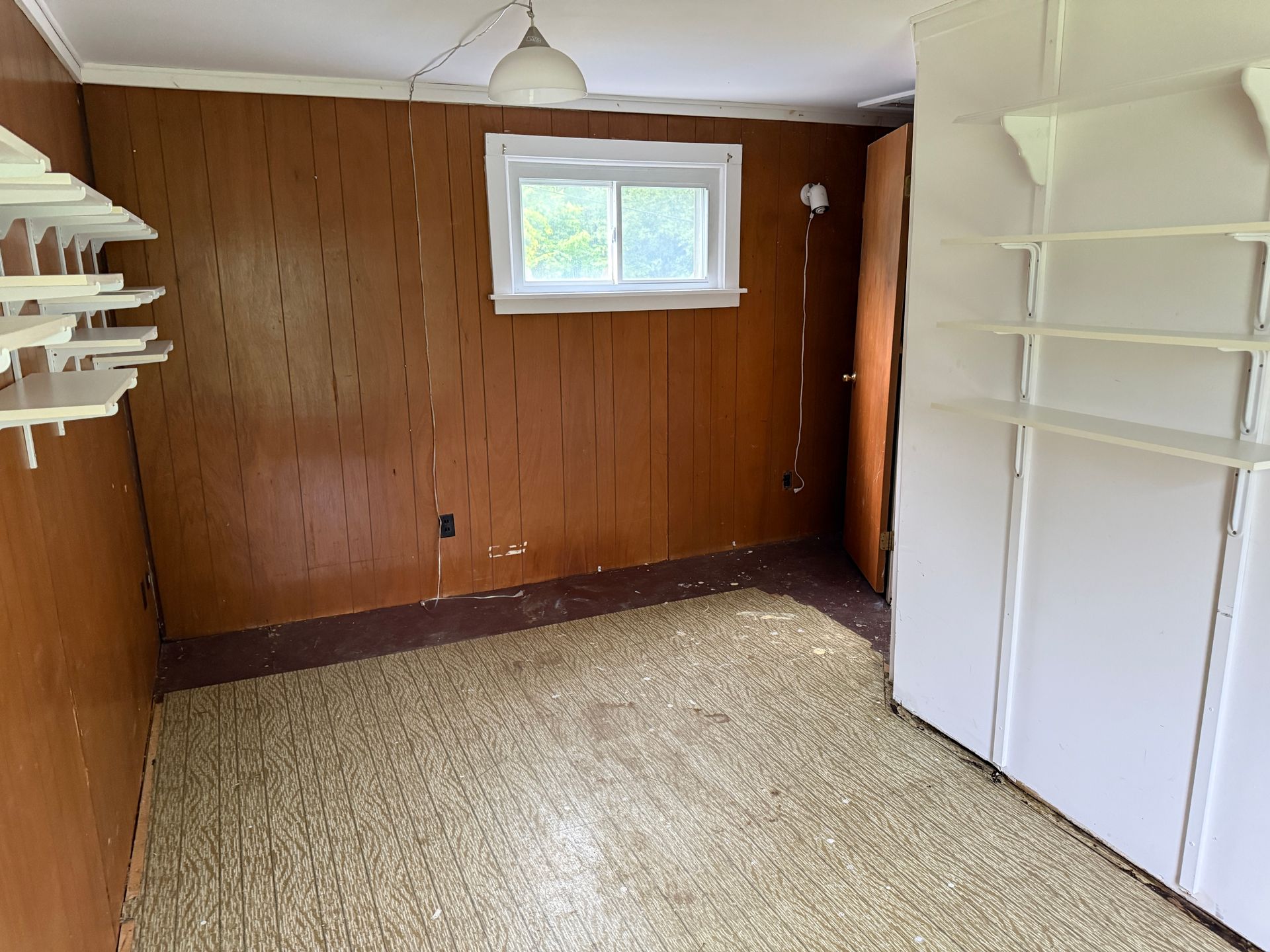 Empty beige-carpeted room with wood-paneled walls, small window, and white built-in shelves and cabinets