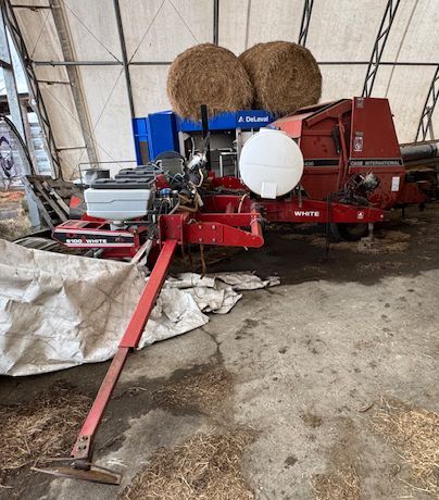 Red farm baler inside a barn, with hay bales and a white wrapped bale nearby.