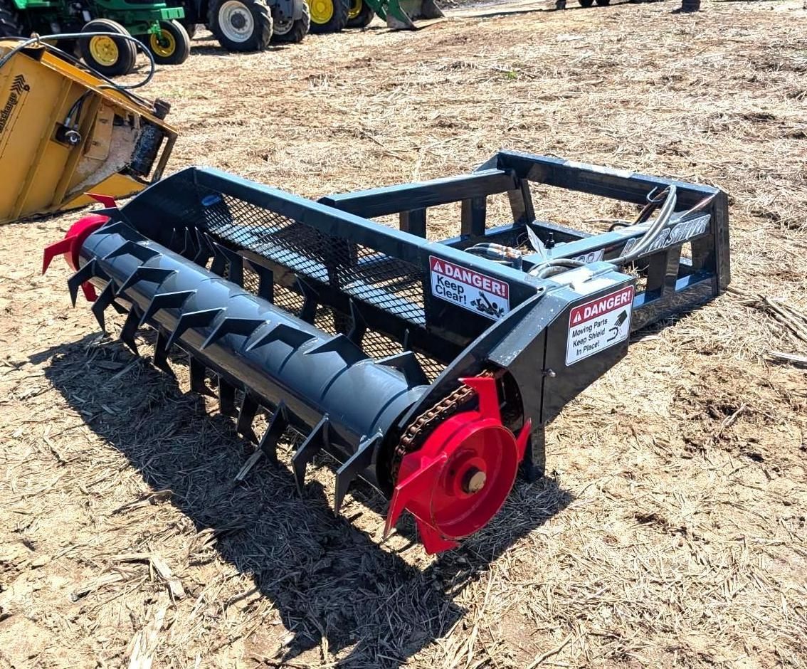 Black rotary tiller attachment with red end caps sitting on field, next to a tractor.
