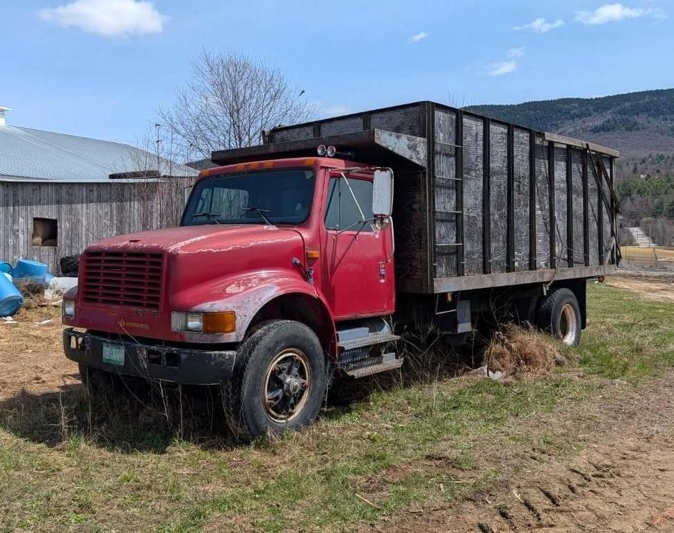 Red dump truck parked on a grassy farm field beside a barn under a blue sky