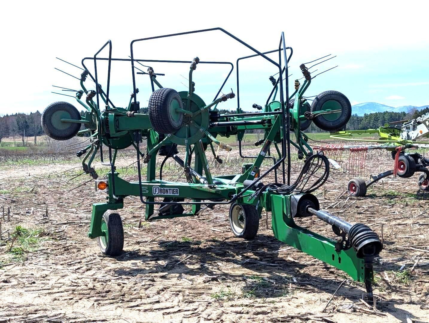 Green agricultural hay rake parked in a field, with multiple wheels and folded arms.