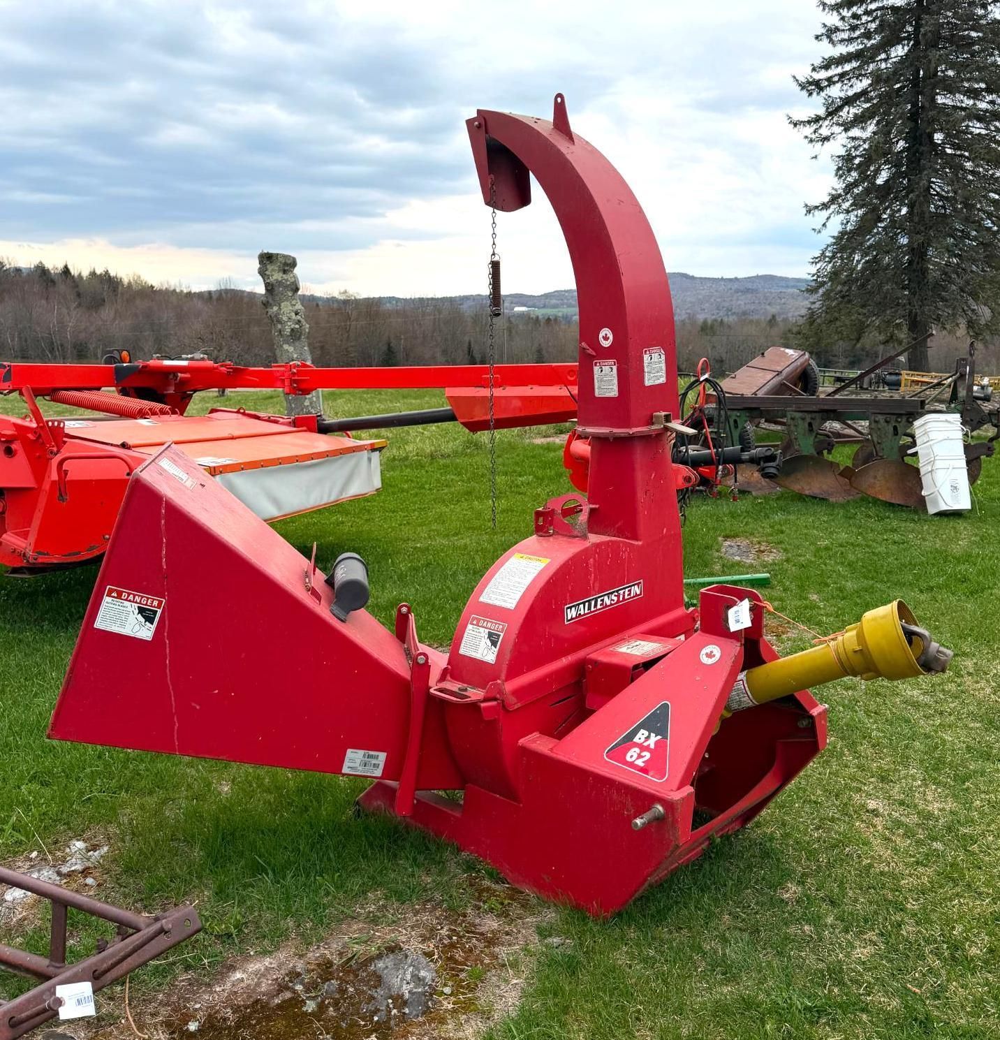 Red agricultural hay processor with yellow PTO shaft sitting on grass in a farmyard.