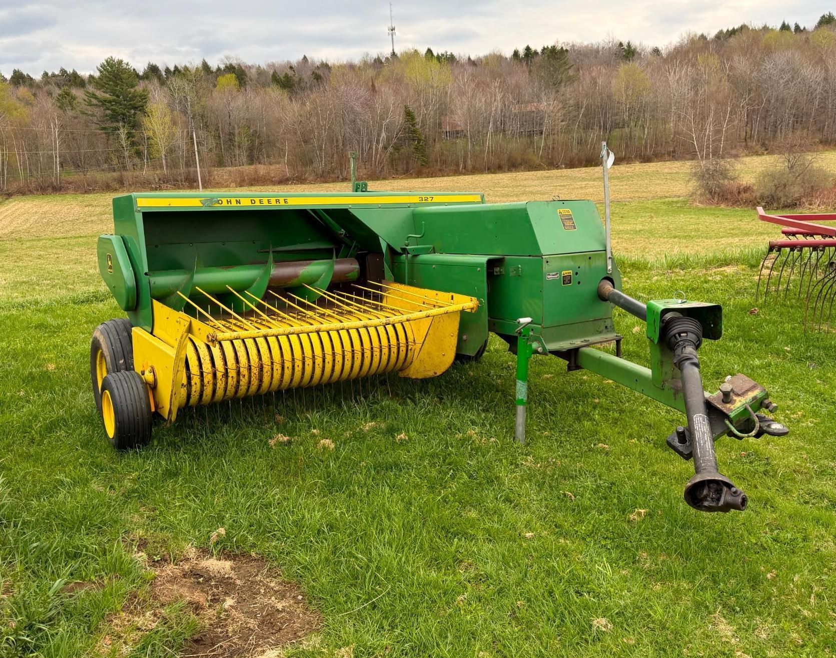 Green hay baler with yellow pickup reel sitting on a grassy field, attached to a tractor hitch